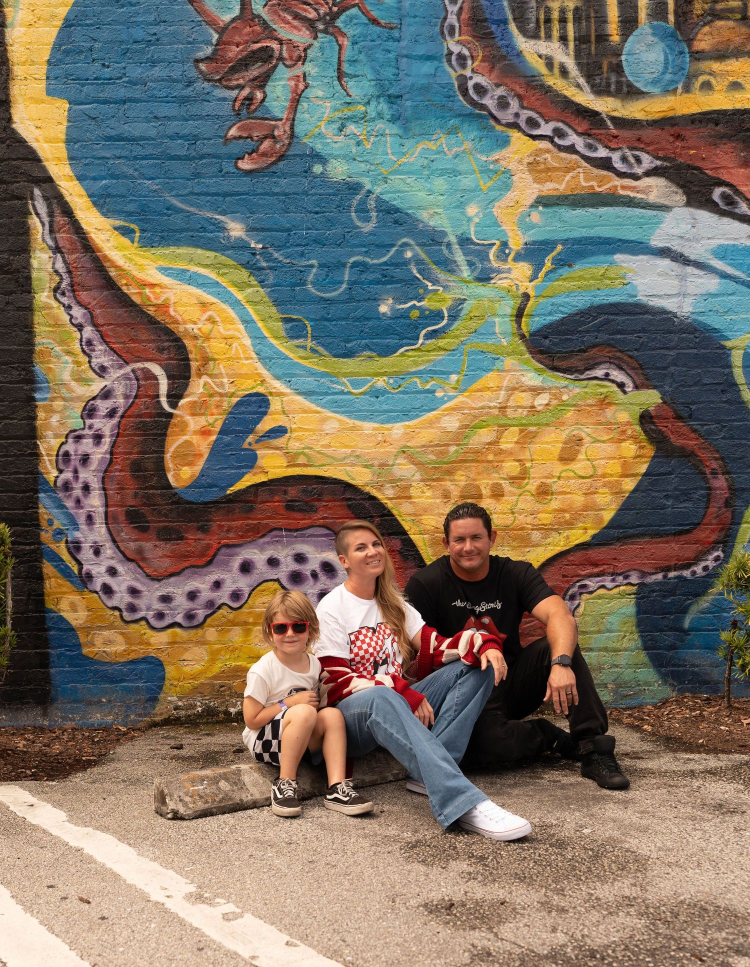 Family of three sitting in front of a colorful octopus street mural on a brick wall outdoors.