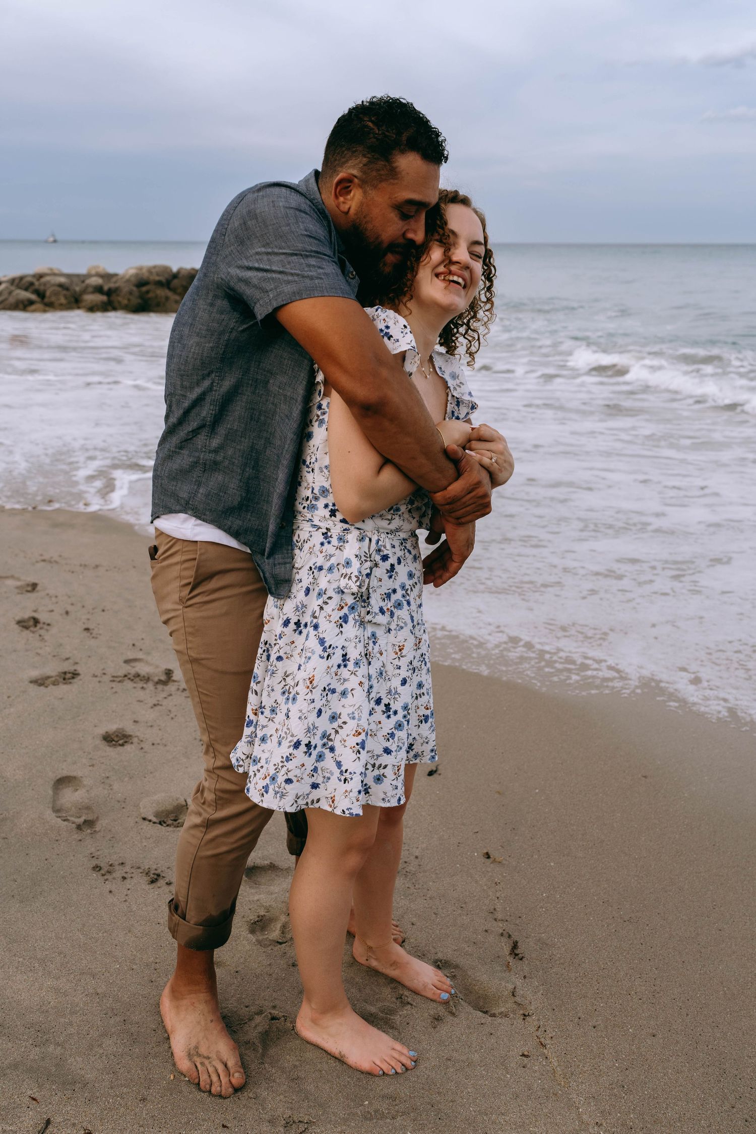 A couple embraces barefoot on a sandy beach, the man hugging the woman from behind as waves crash nearby on Worth Avenue Palm beach.