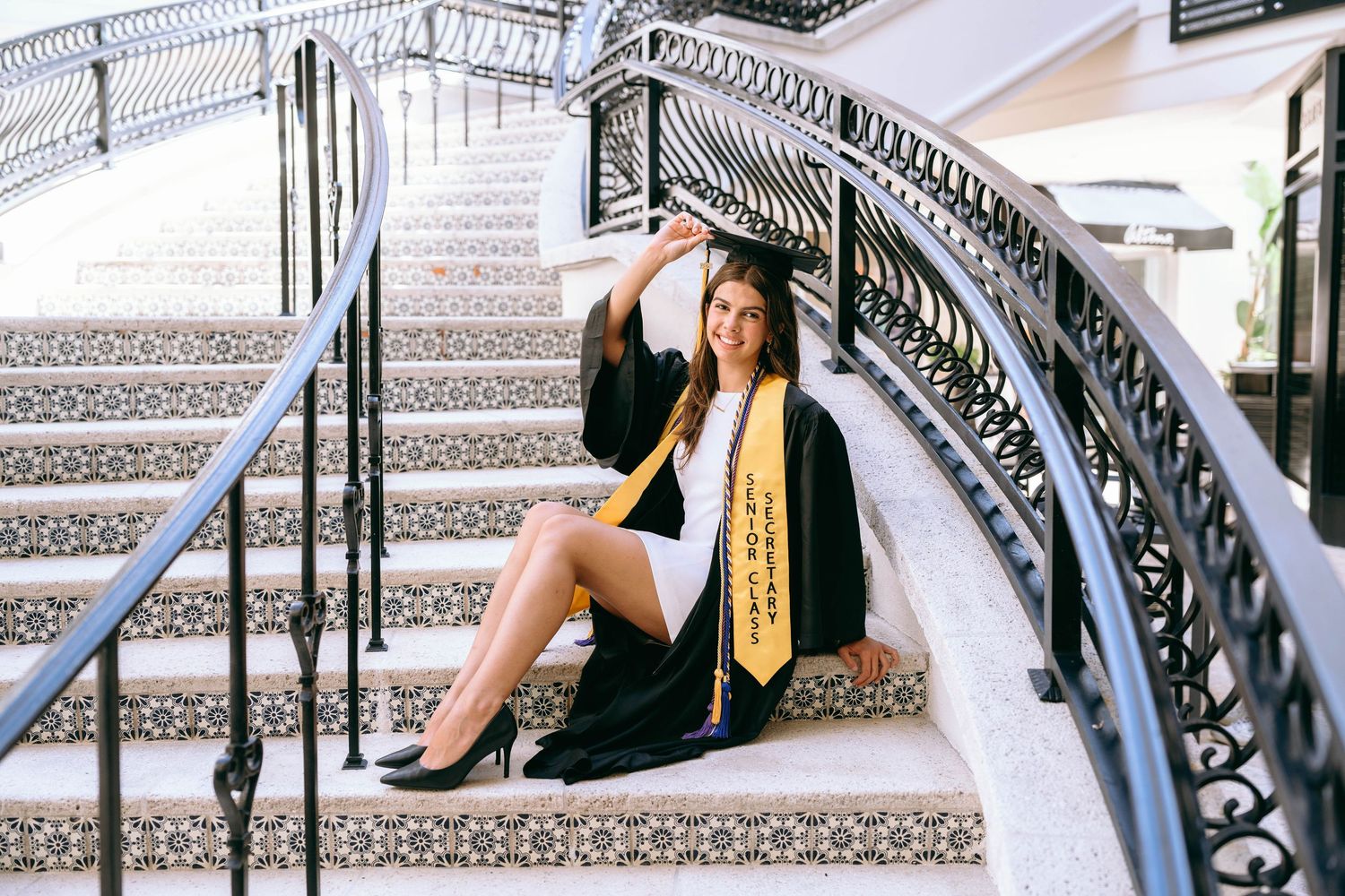 A smiling graduate in black cap and gown with gold sash sits on ornate tiled stairs for a graduation portrait on Worth Avenue Palm beach.