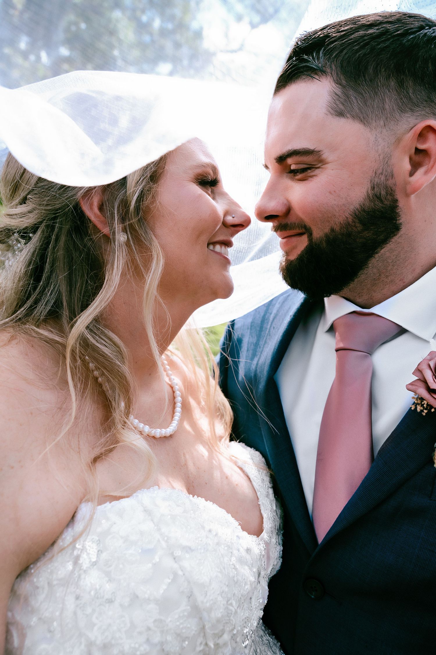 Smiling bride and bearded groom share romantic moment under veil, groom wearing navy suit with pink tie.