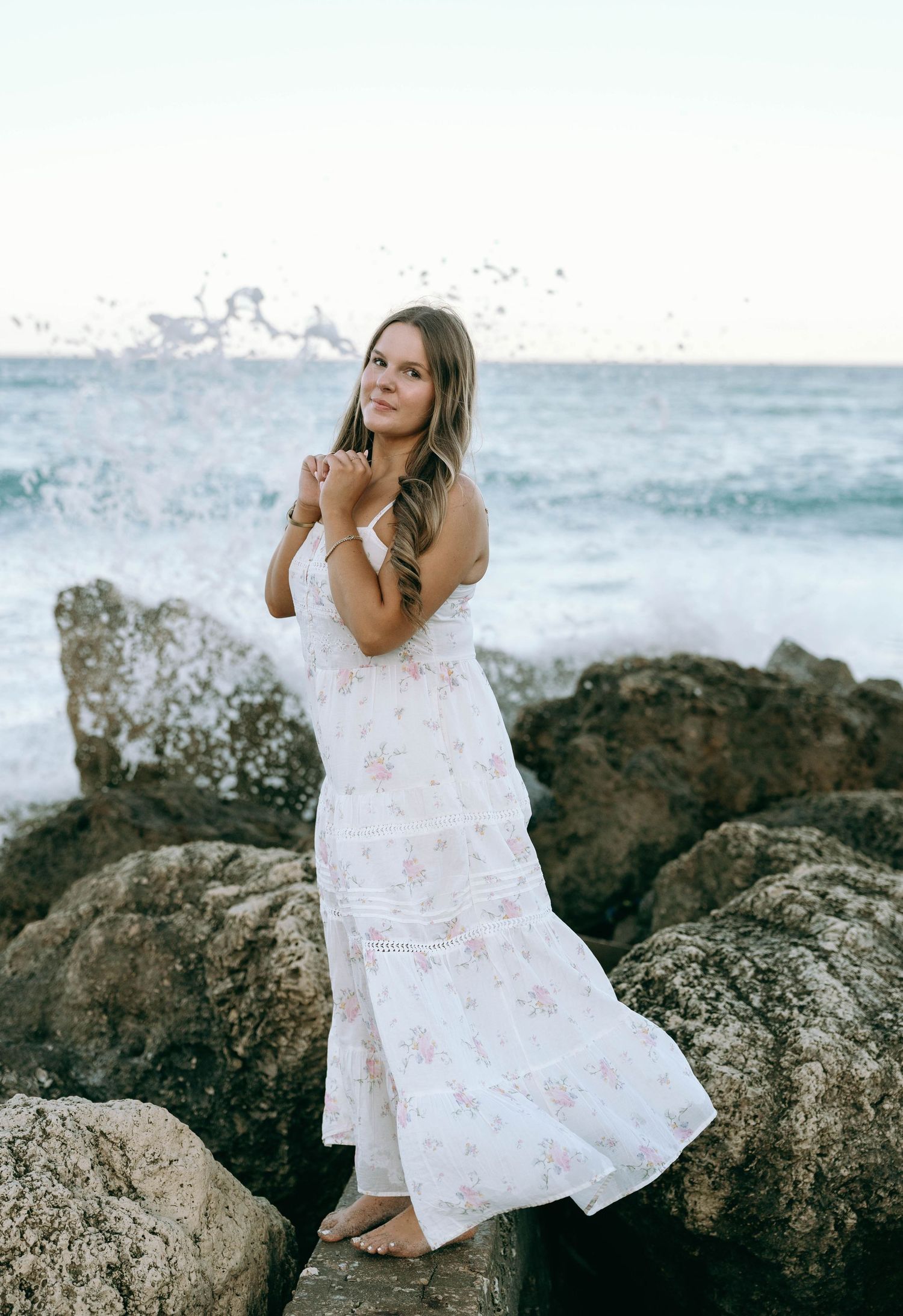 Bride in floral white maxi dress stands on rocky shore with ocean waves crashing behind her. Worth Avenue, Palm Beach, FL