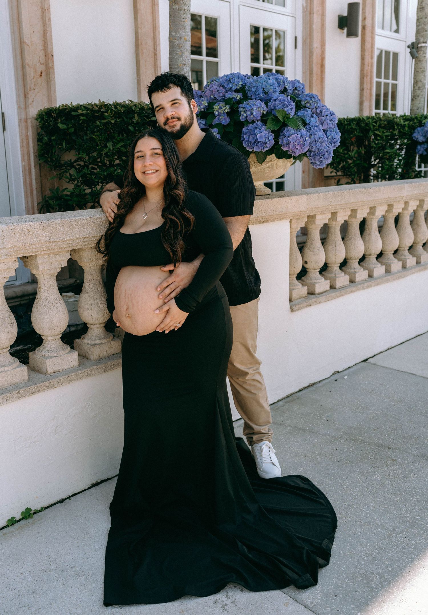 Smiling pregnant couple in black posing on elegant balcony with purple hydrangeas. Worth Avenue, Palm Beach, FL
