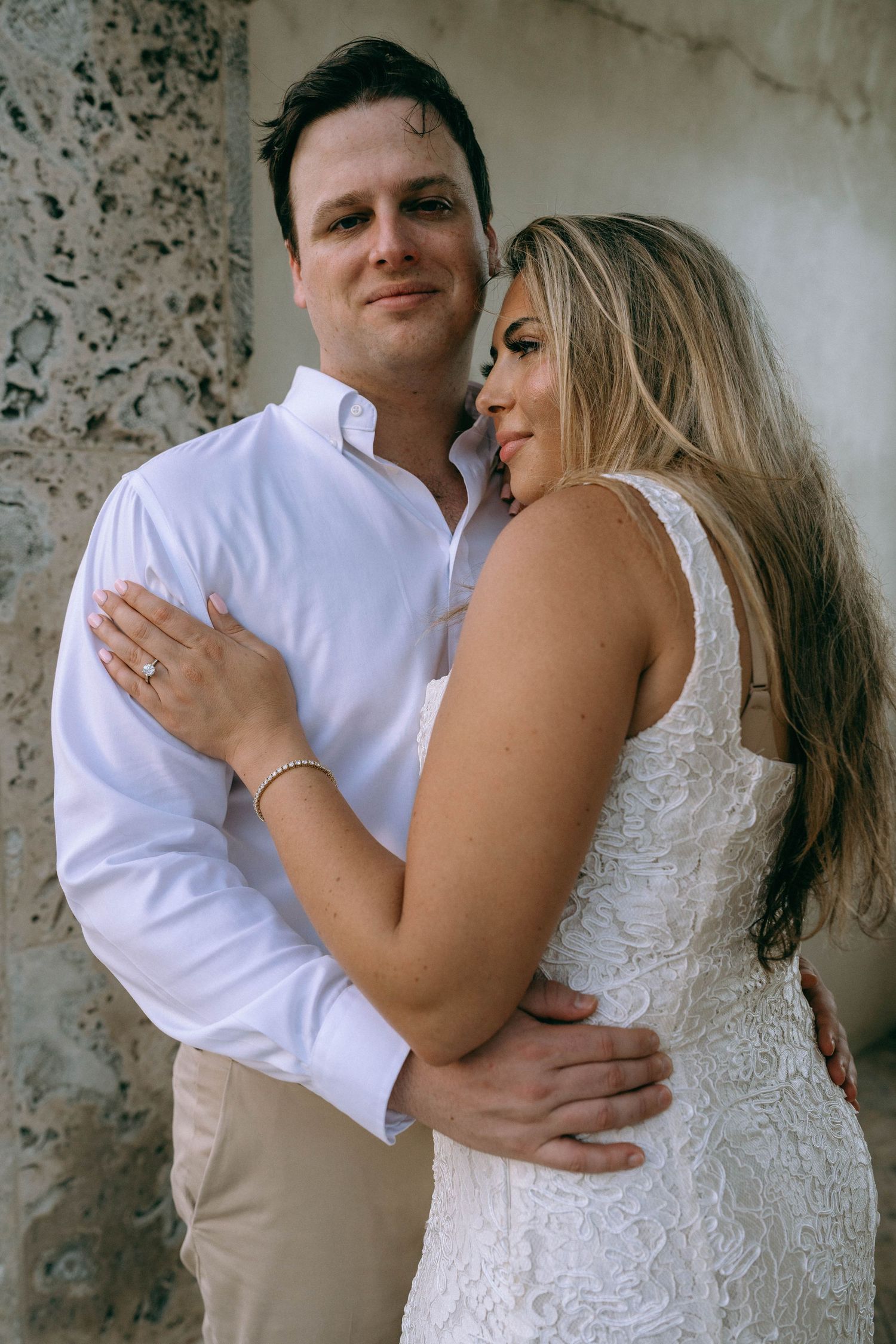 Man in white shirt tenderly holds woman in white lace dress close, posing against a stone wall.
