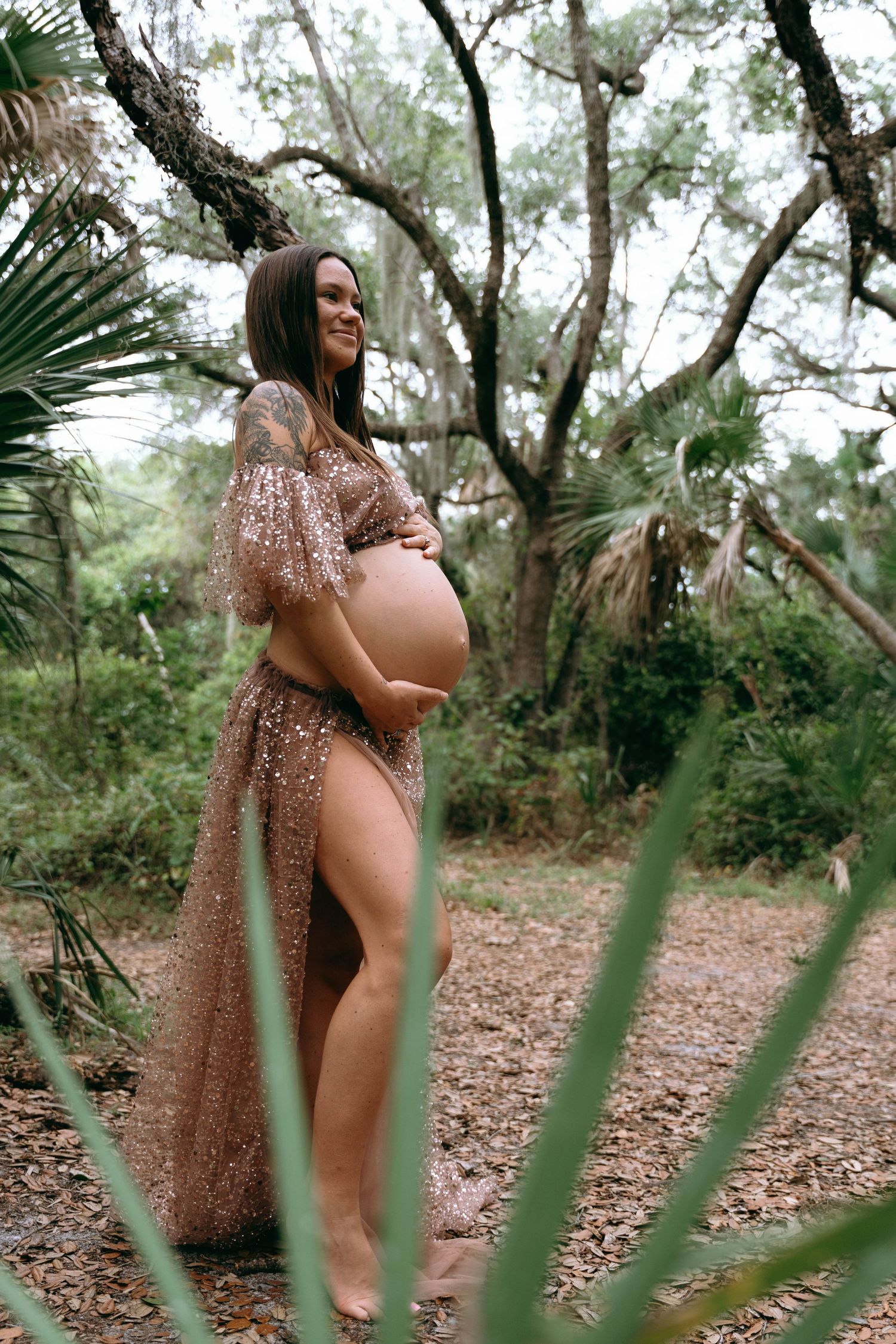 A maternity photoshoot in a natural wooded setting with palm fronds and trees.