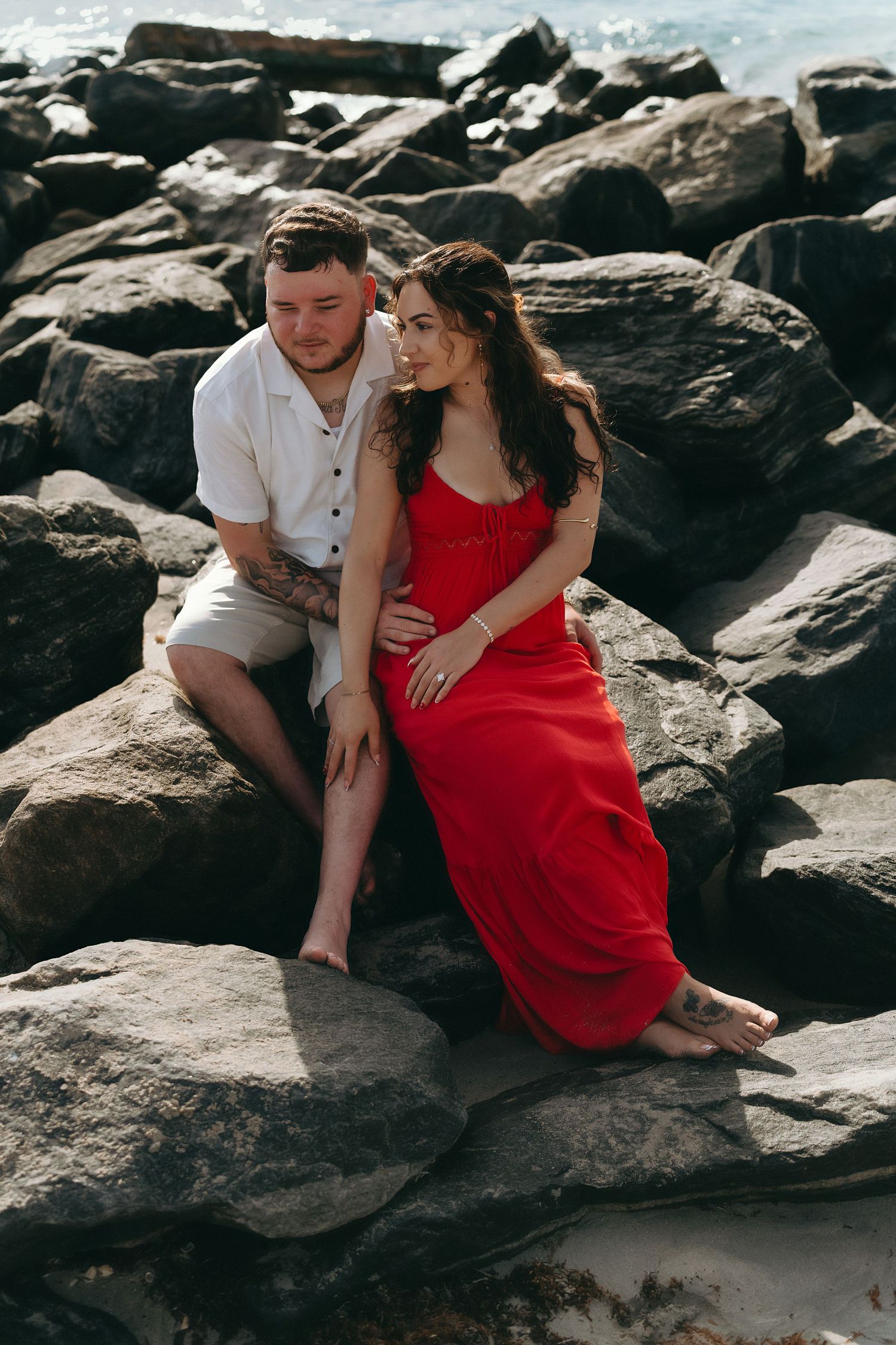 Couple in white and red outfits embrace on rocky beach during sunset.