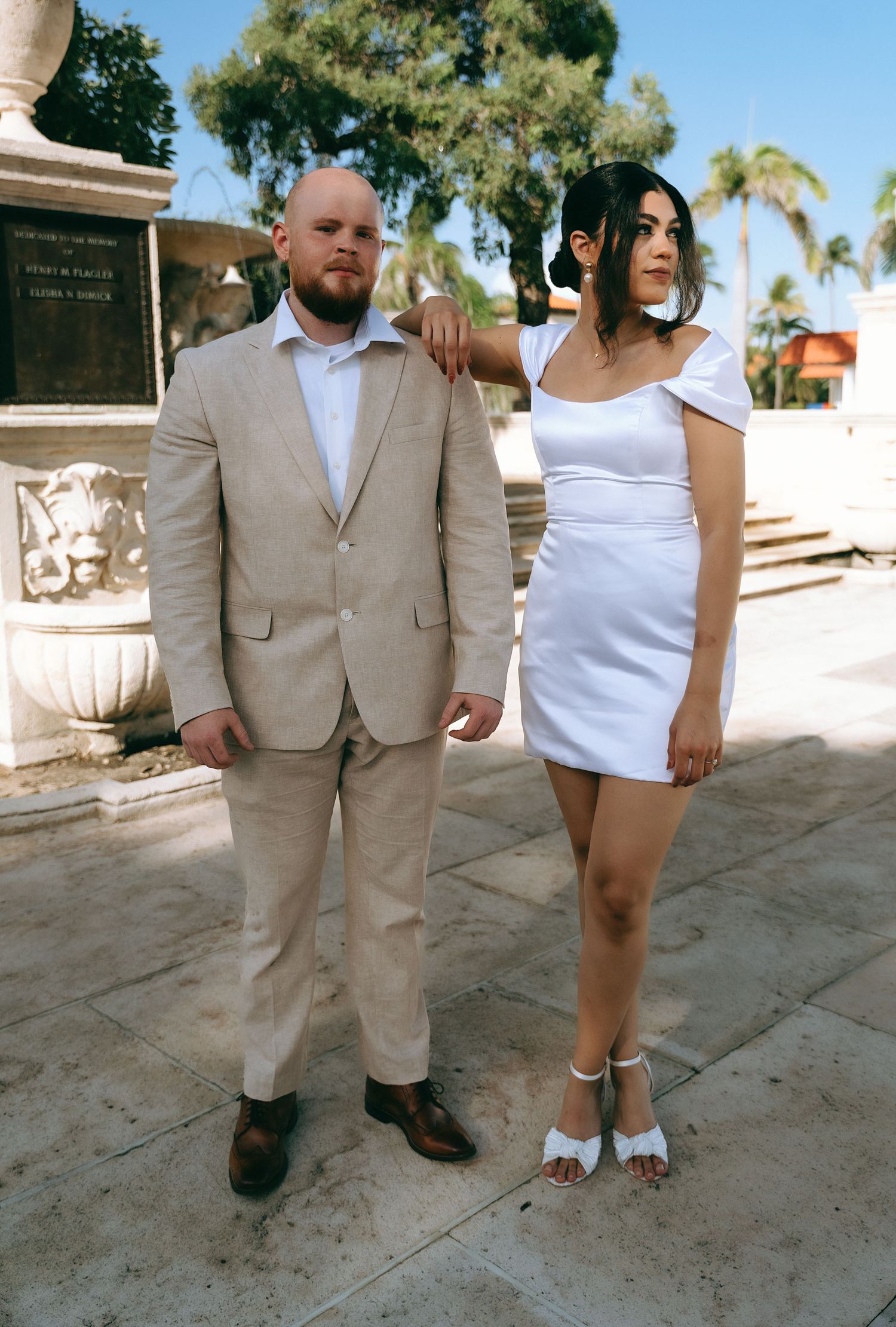 Couple in summer wedding attire posing on stone path with palm trees.