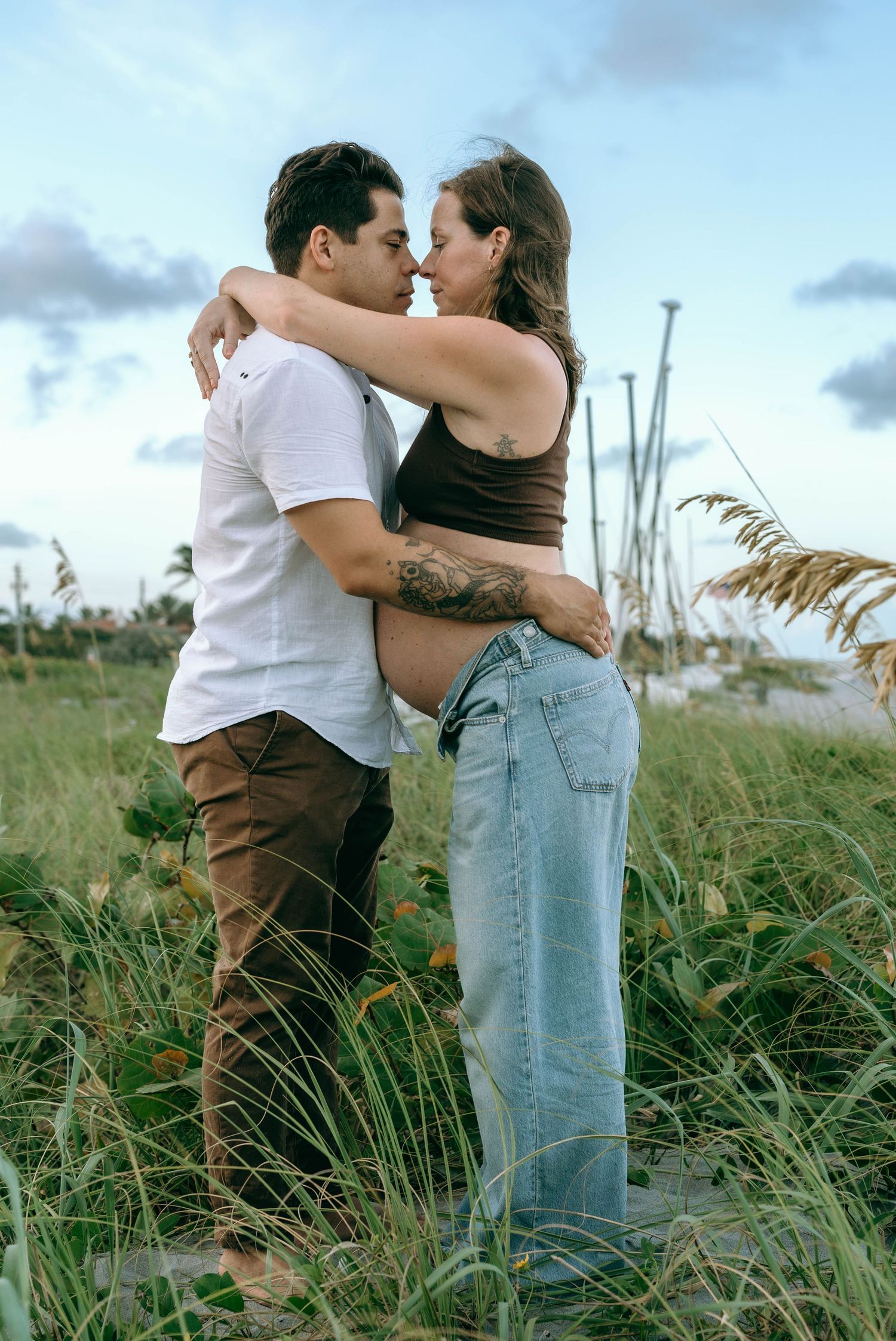 Couple embracing in tall grass near the beach, pregnant woman in jeans and crop top with tattoos, romantic outdoor maternity photo.