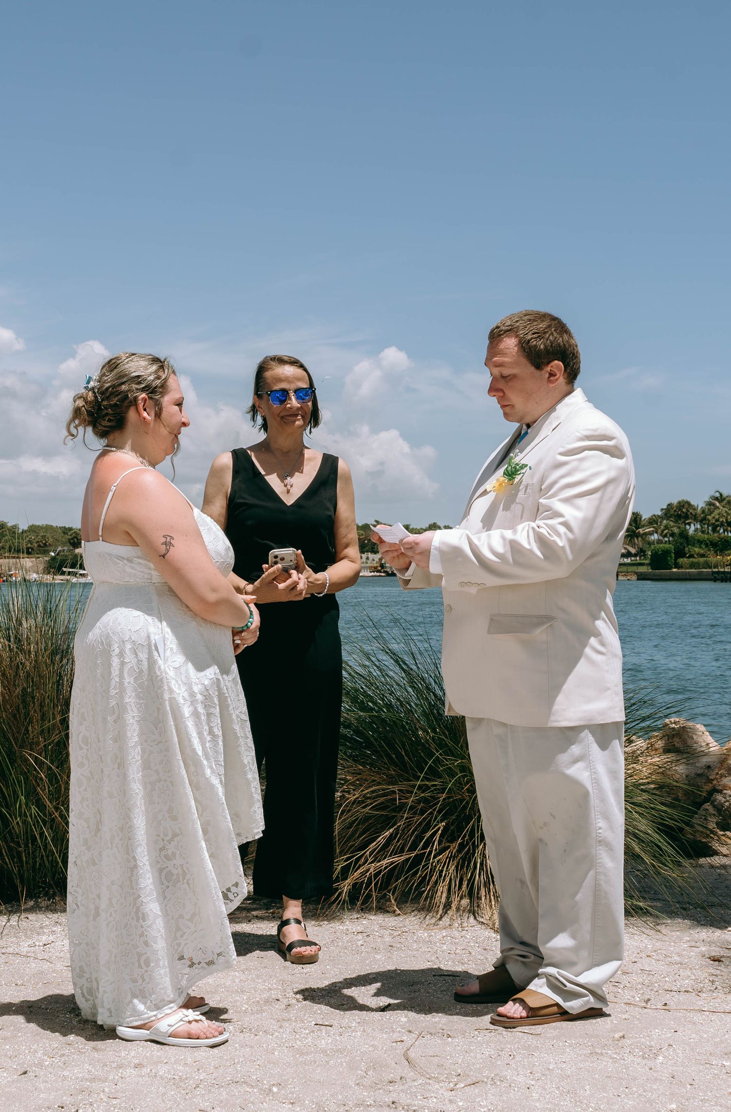 Outdoor wedding ceremony by the water with bride in white dress, groom in white suit, and officiant in black dress.