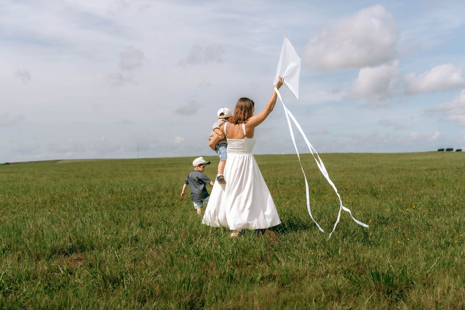 Woman in white dress flying a kite with two children in a green field under a cloudy sky.