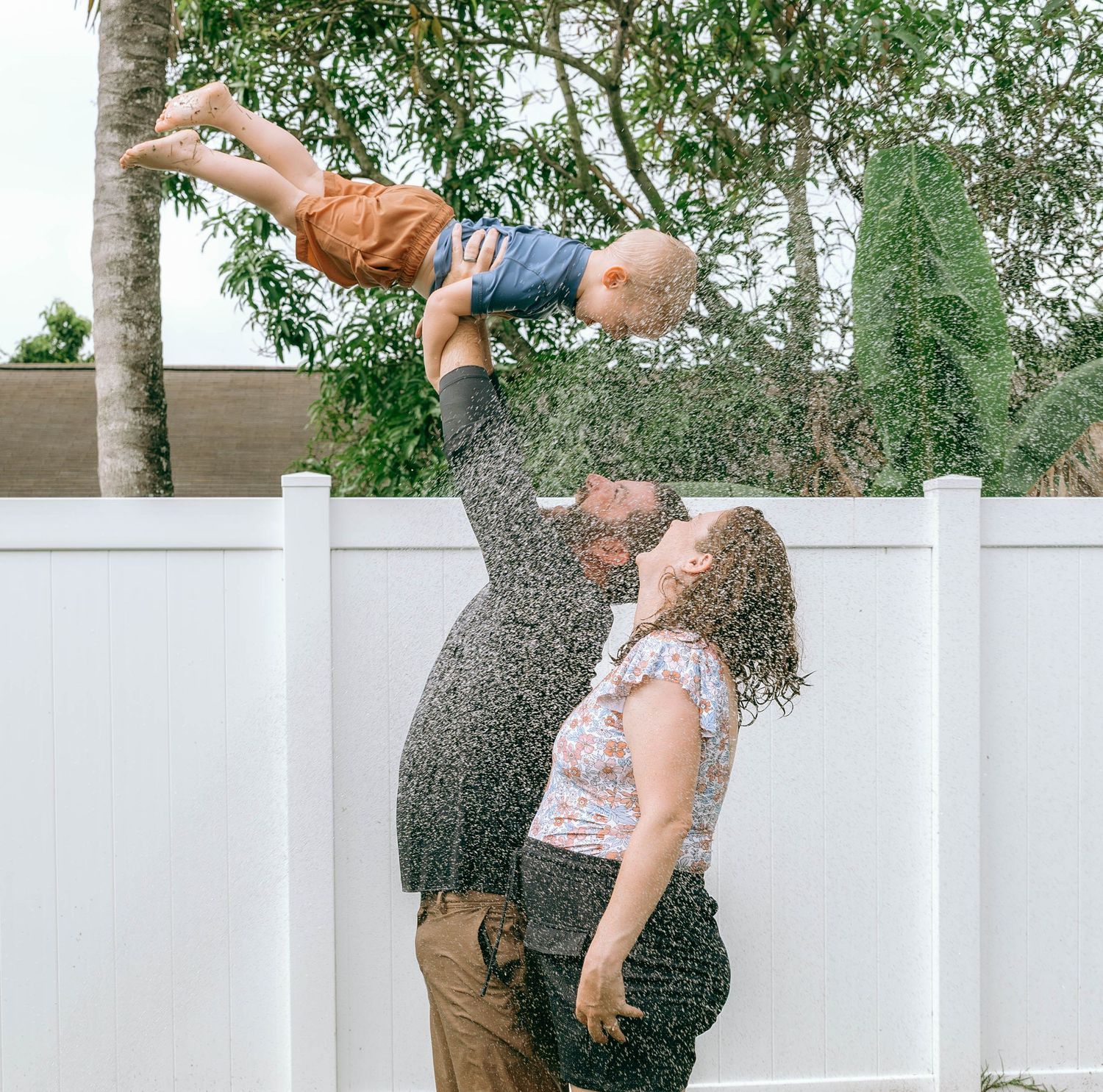 Father lifts toddler high above while mother watches lovingly in sunny backyard.