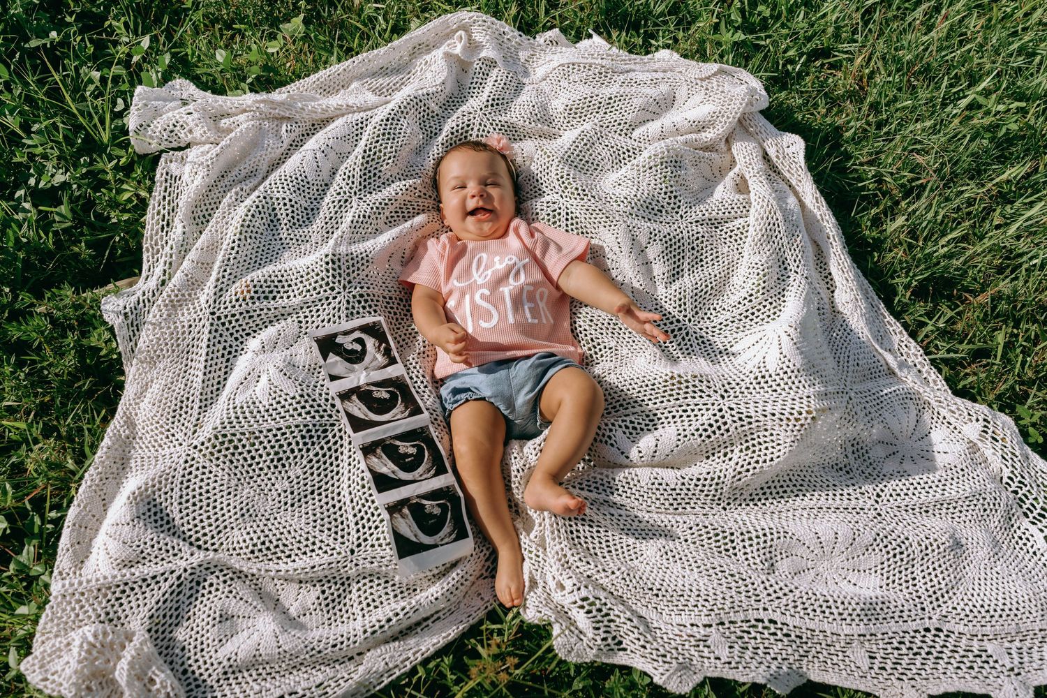 Baby girl in pink top and denim shorts laughing on white lace blanket on green grass outdoors.
