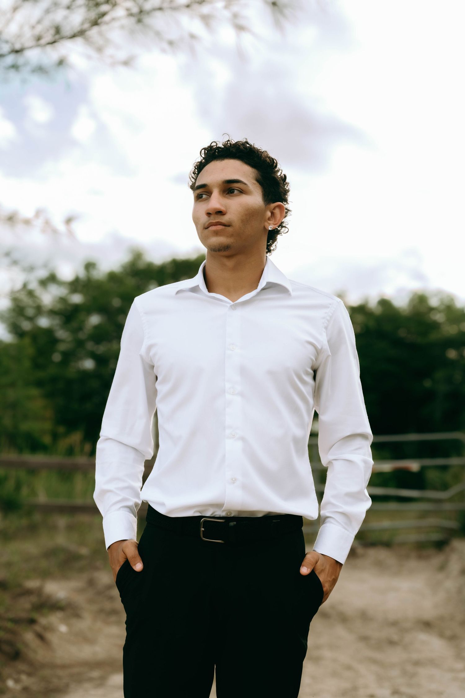 Young man in white dress shirt and black pants stands outdoors with hands in pockets, looking away confidently. Senior Photos at Colt Farms