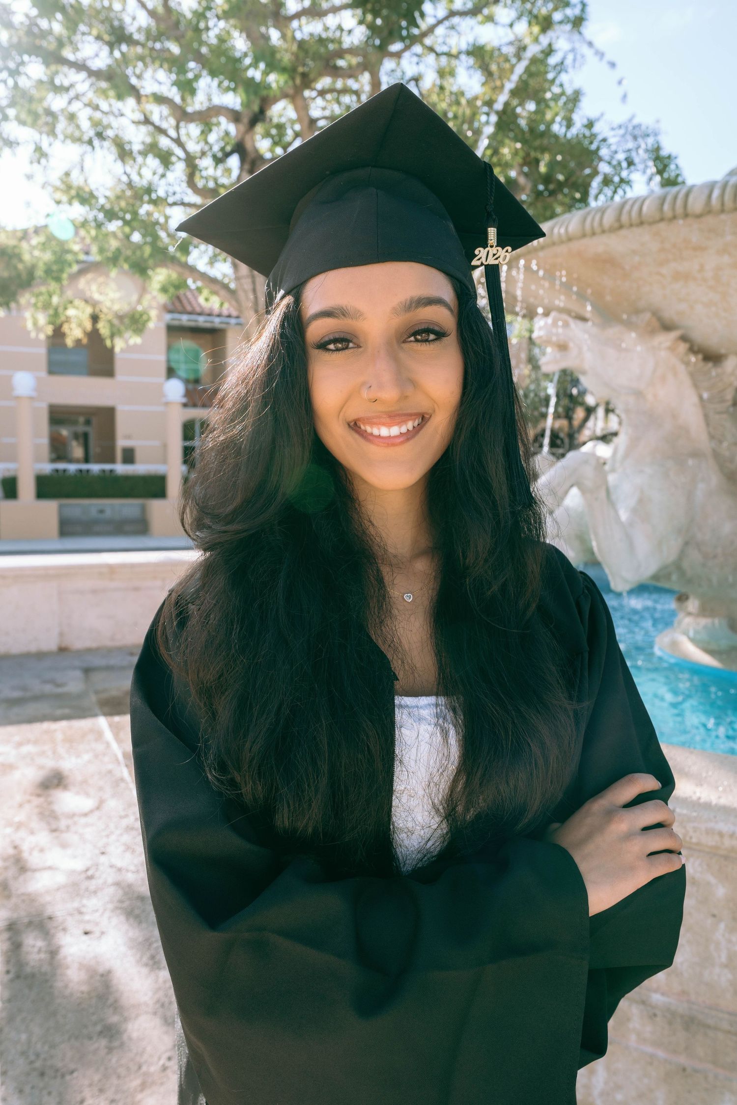 Smiling graduate in black cap and gown with arms crossed standing outdoors near a fountain on Worth Avenue Palm beach.