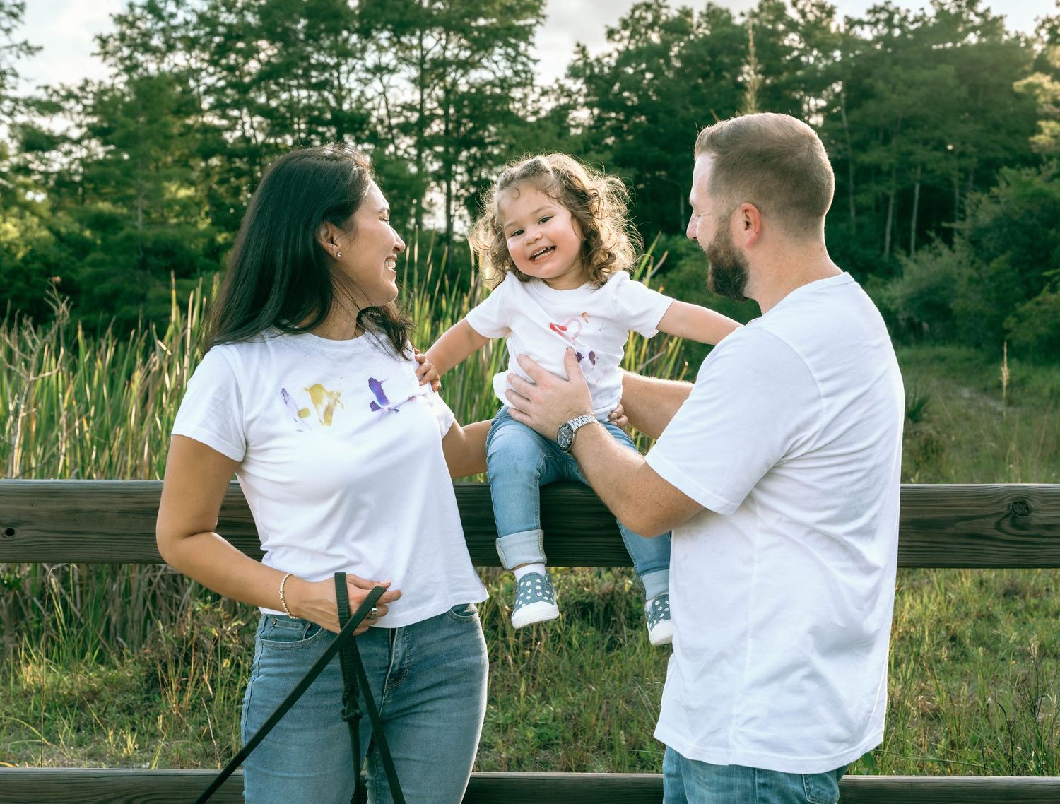 A happy family of three in matching white t-shirts poses outdoors near a wooden fence with lush greenery.