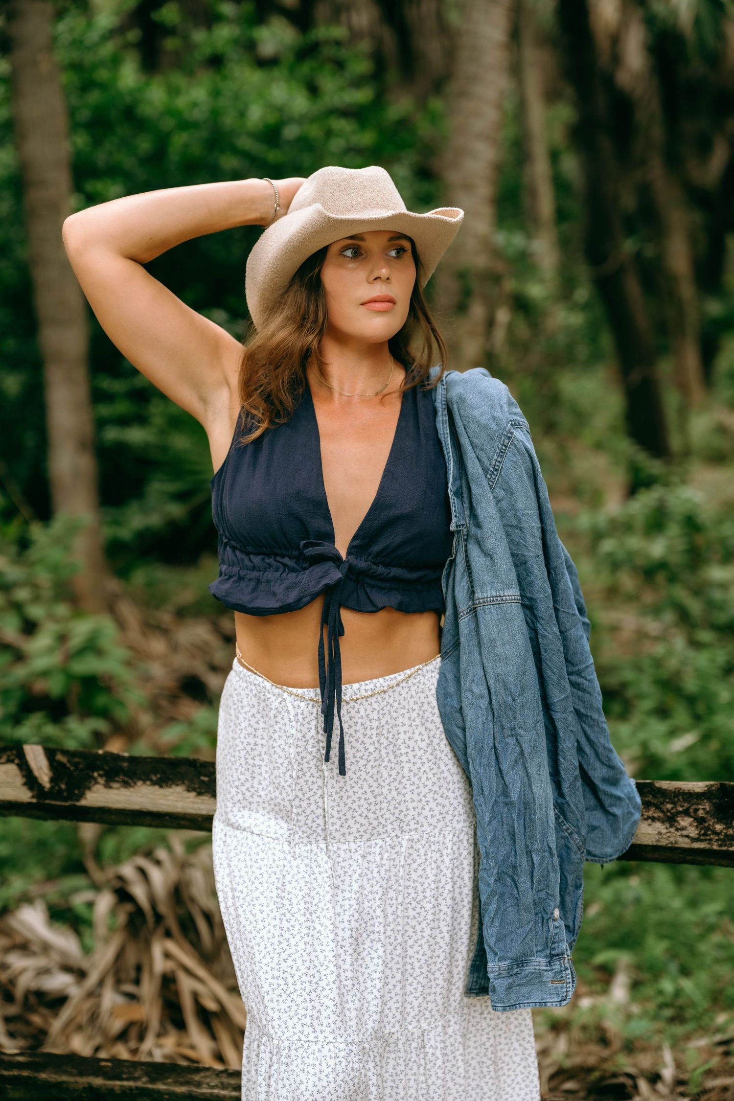 Woman in cowboy hat, black crop top, white skirt, and denim jacket posing outdoors in a lush green forest setting.