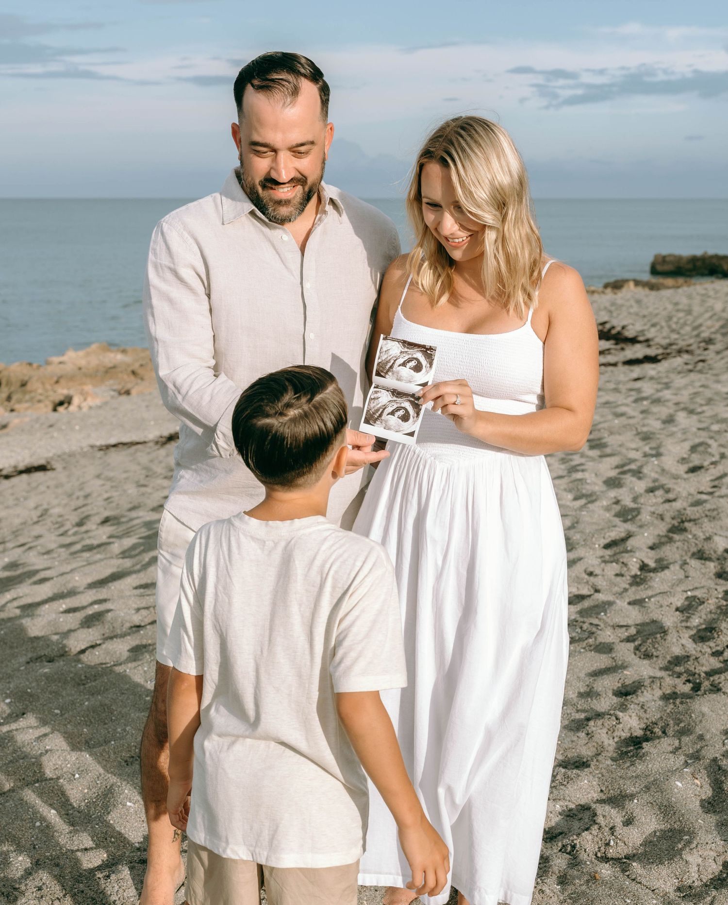 Family on beach sharing ultrasound photos, parents smiling at child in white outfits with ocean backdrop.