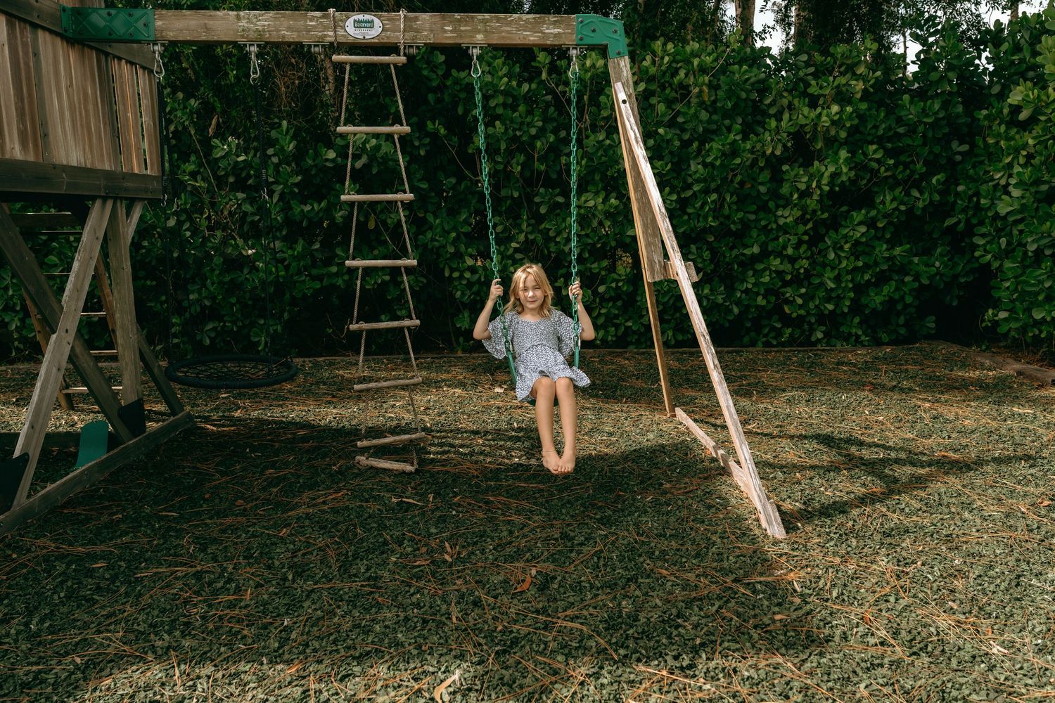 Young girl with blonde hair smiling while swinging on a wooden swing set in a lush green backyard.