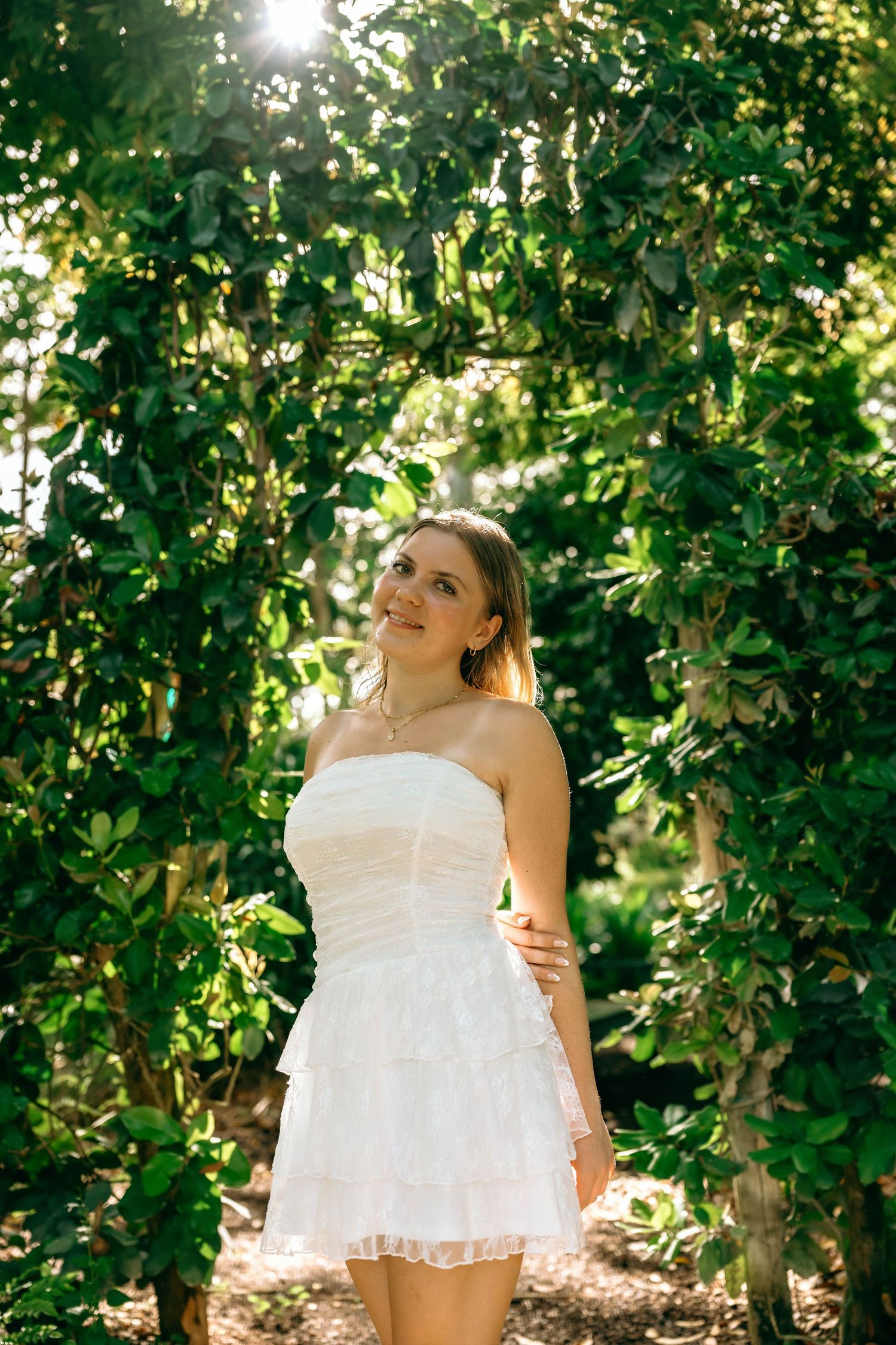 Smiling young woman in white strapless mini dress standing under lush green garden archway in sunlight.
