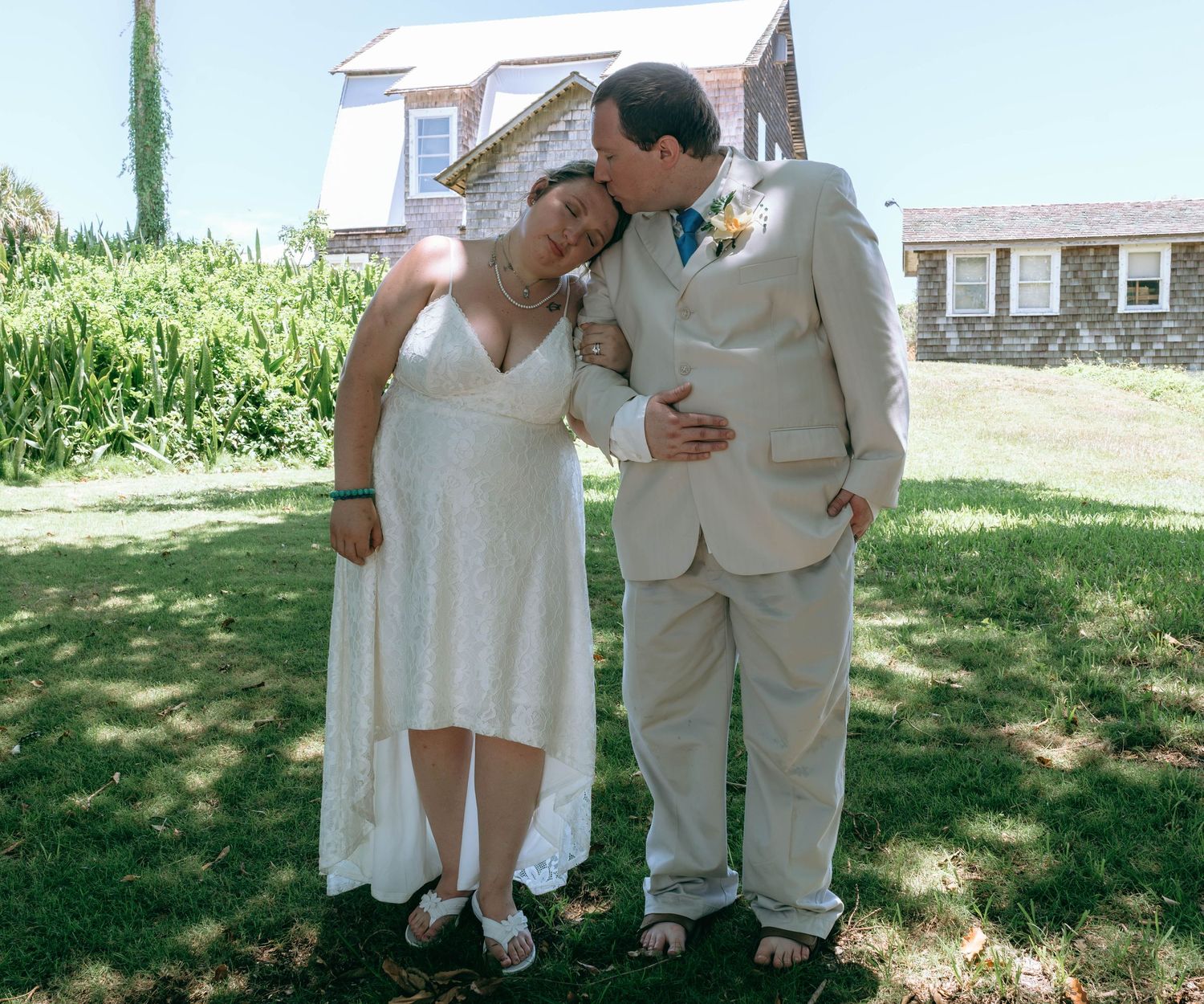 Bride and groom share a tender moment in white wedding attire on a sunny green lawn outside a stone house.