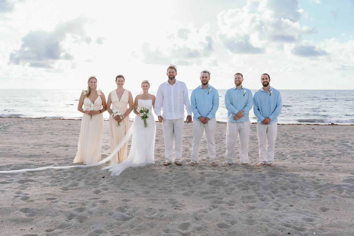 Wedding party lined up on beach in white and light blue attire.