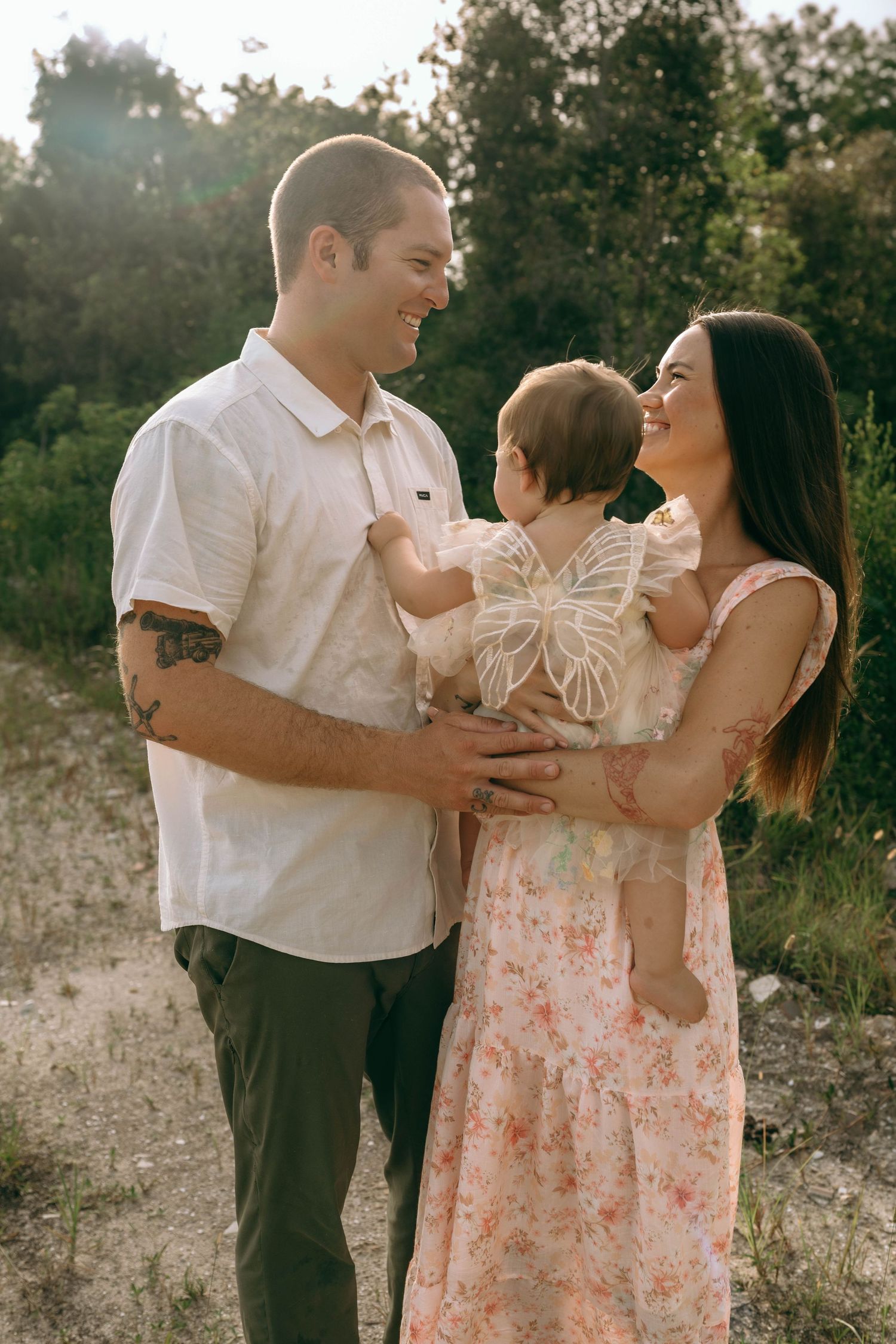 A smiling couple holds their baby girl in a sunny outdoor setting, the woman wearing a floral dress and man in a white shirt.