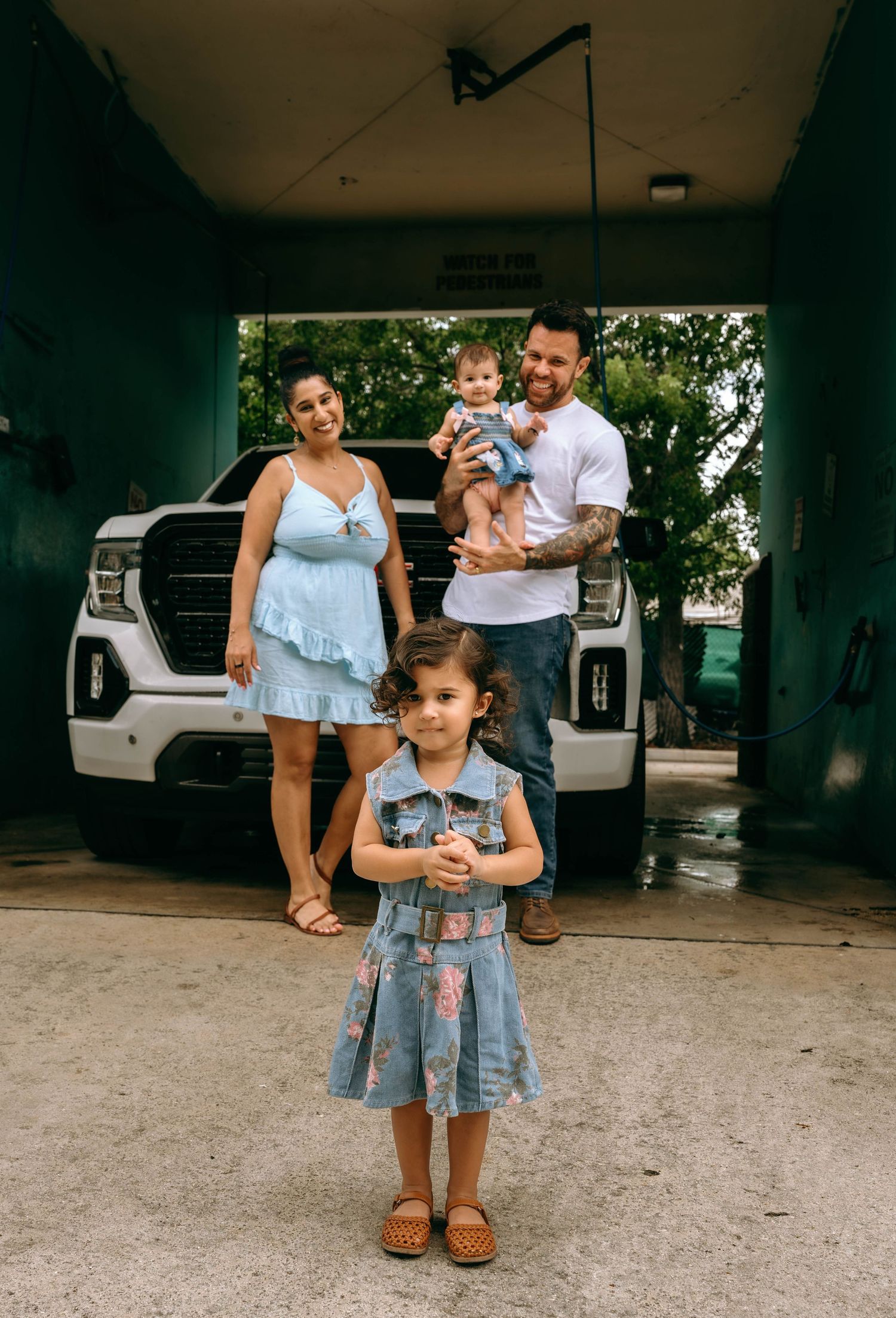 Family of four posing in front of white GMC truck, mother in blue dress, father holding baby, young girl in denim dress.