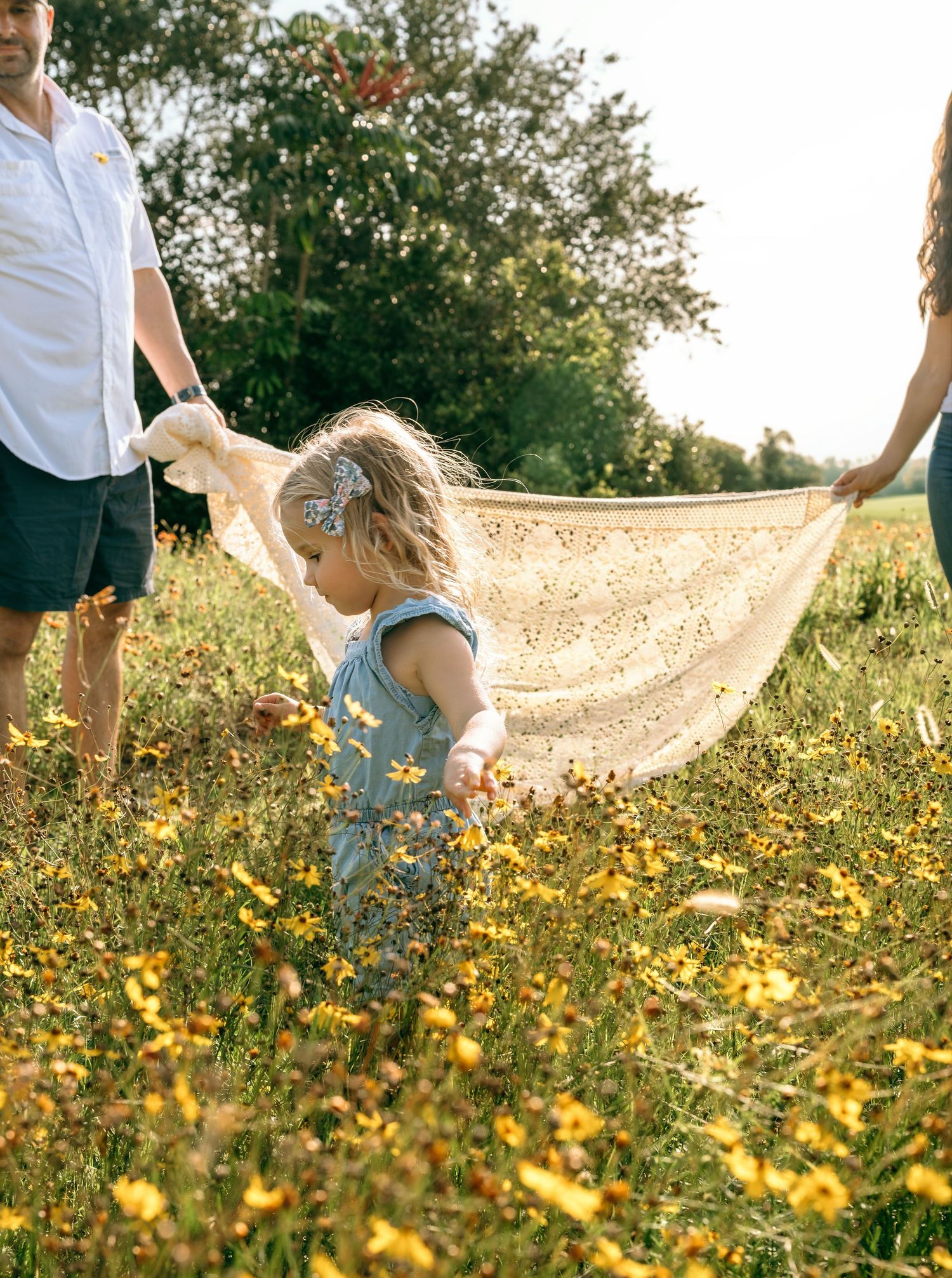 A toddler in denim overalls walks through a sunny wildflower field, held by parents waving a lace blanket.
