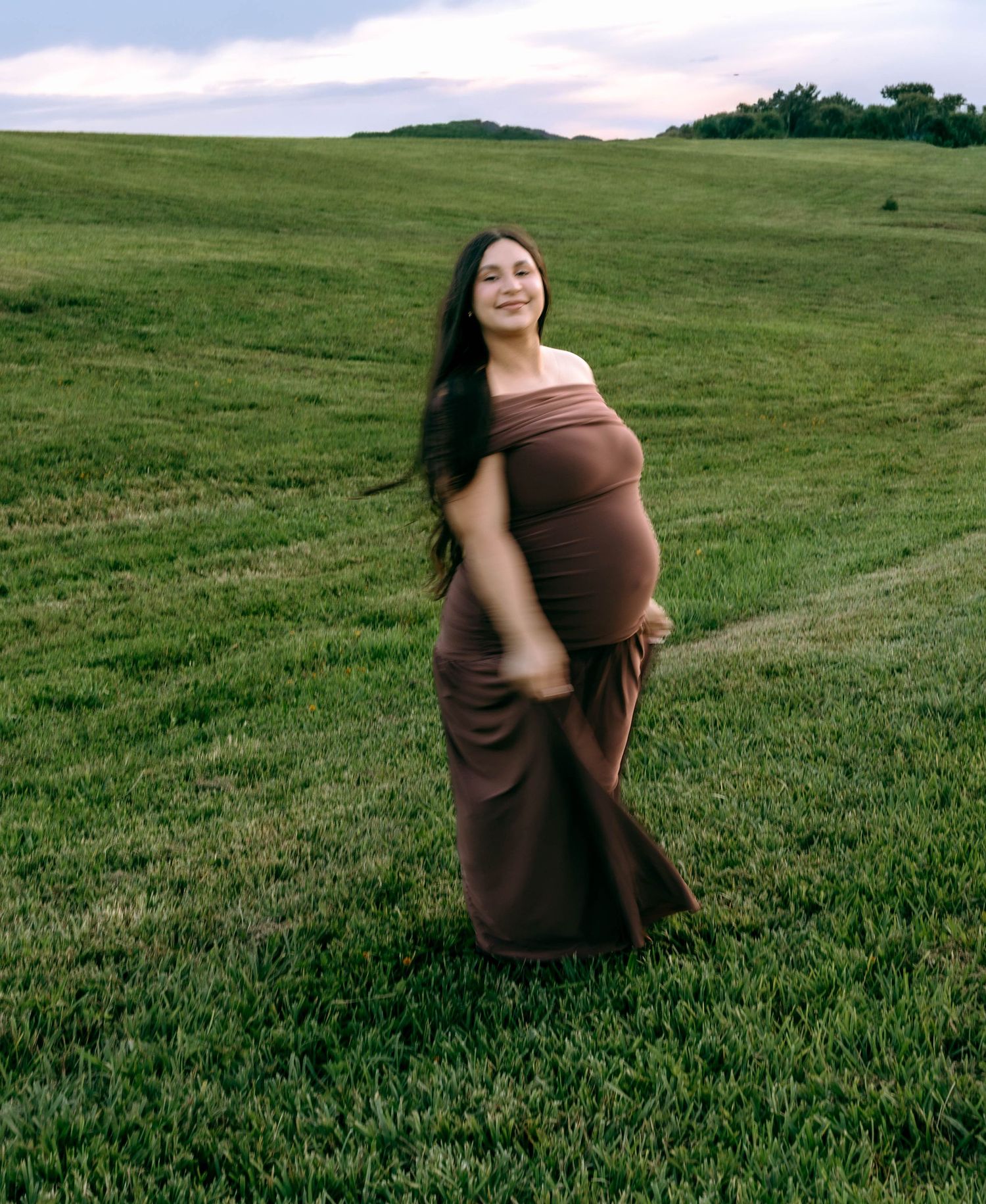Pregnant woman in brown maternity dress standing in green field, smiling with long dark hair flowing.