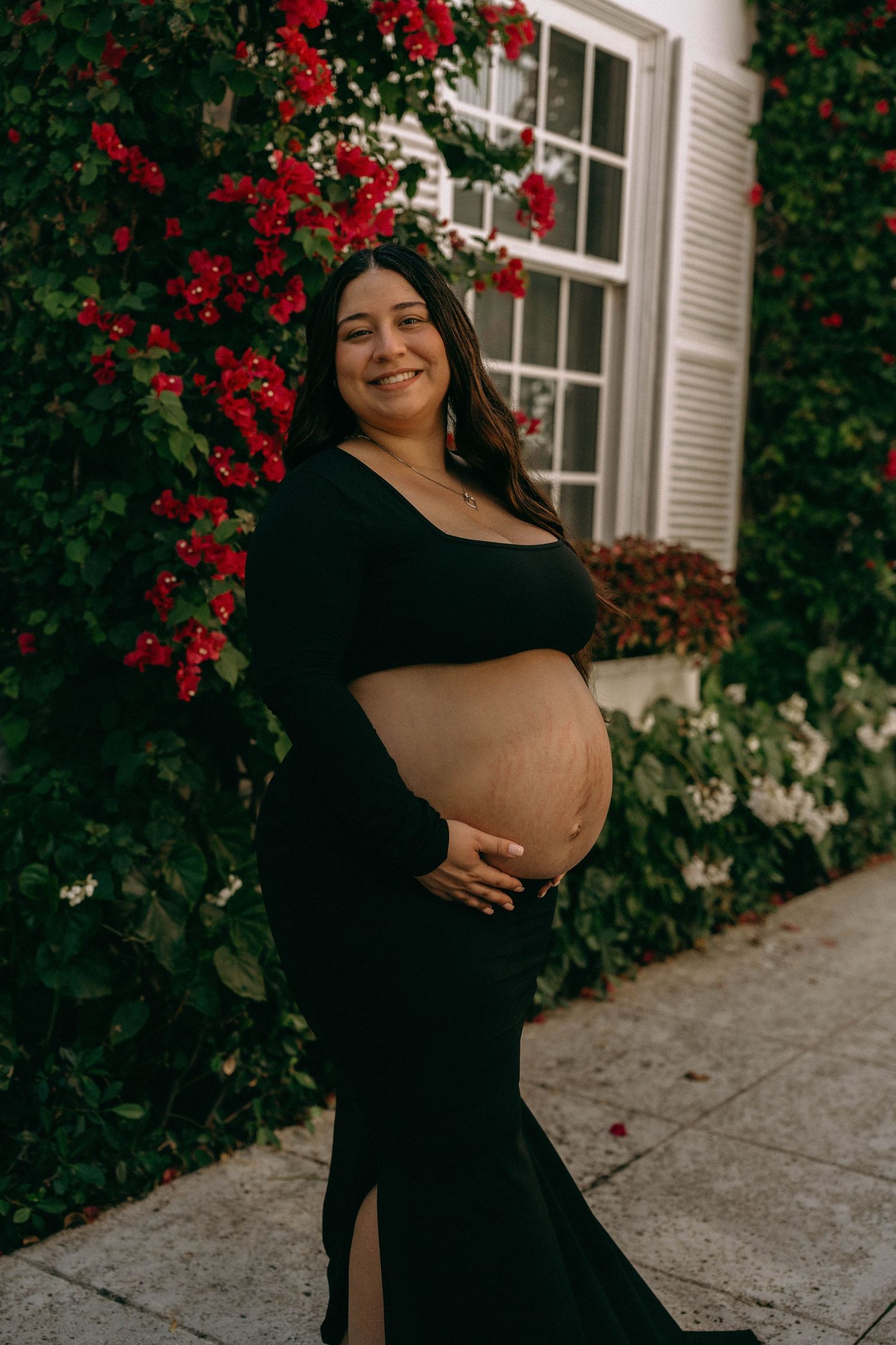 Radiant pregnant woman in black dress cradling baby bump beside lush red bougainvillea wall. Worth Avenue, Palm Beach, FL