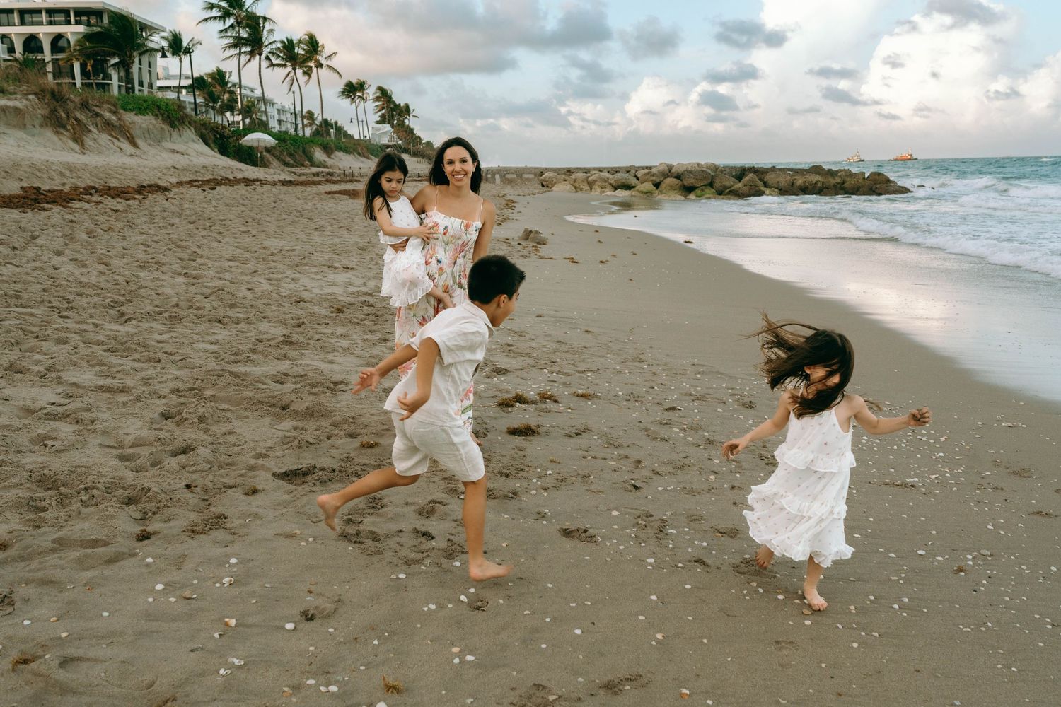 A mother and three children in white outfits play and run together on a sandy beach at dusk. Worth Avenue, Palm Beach, FL
