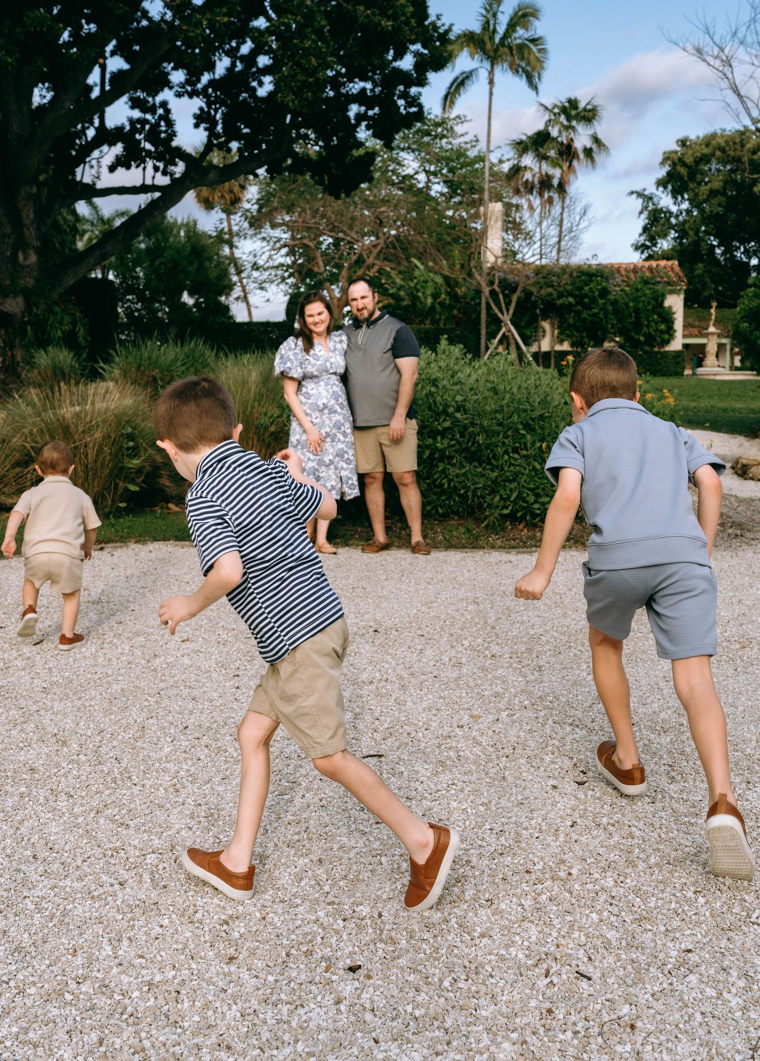 A happy couple watches their three young boys playing outdoors on a gravel path surrounded by lush greenery.  Family photos at Bradley Park