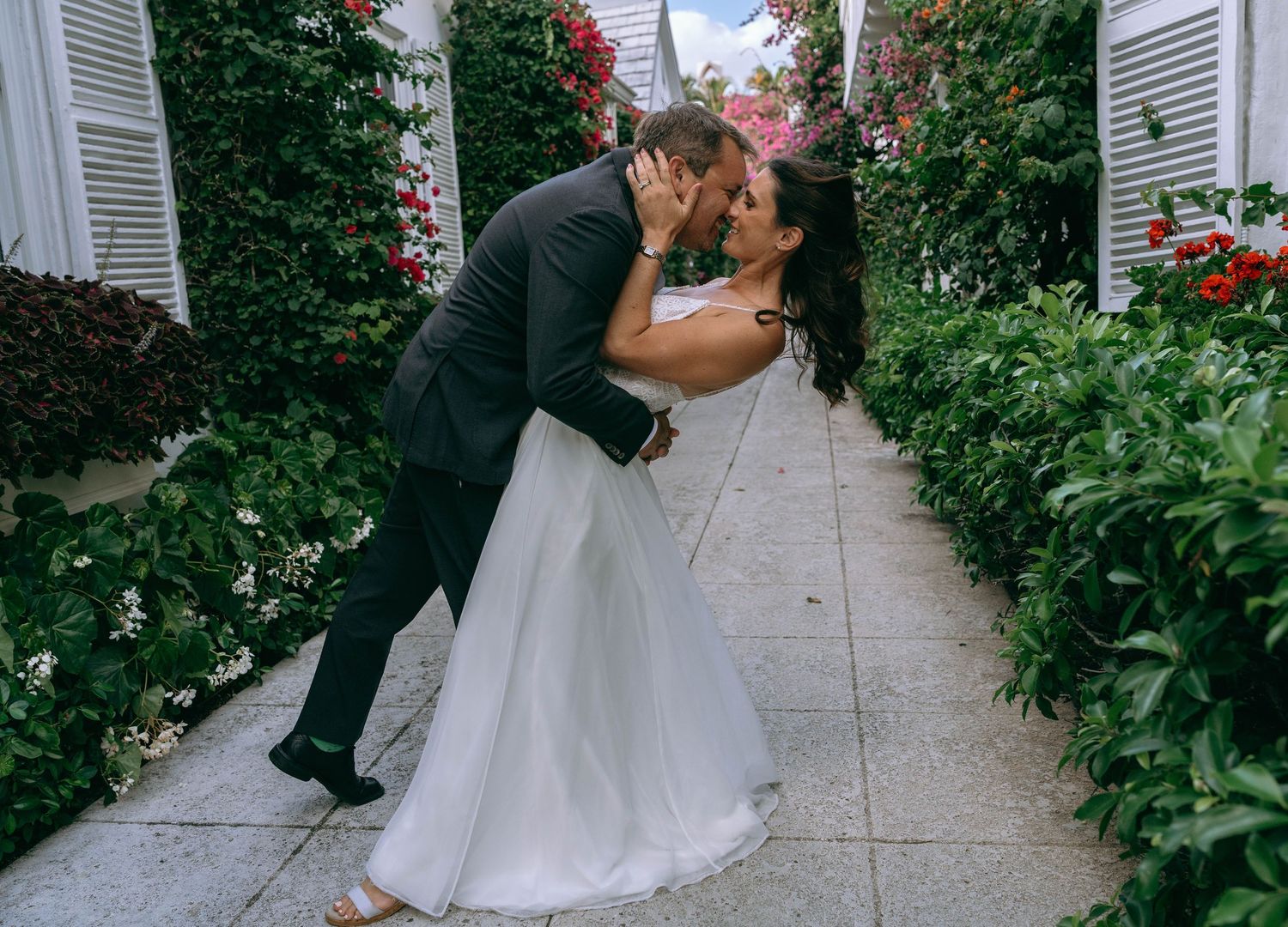 Bride and groom sharing a romantic dip kiss in a lush garden pathway lined with red flowers.