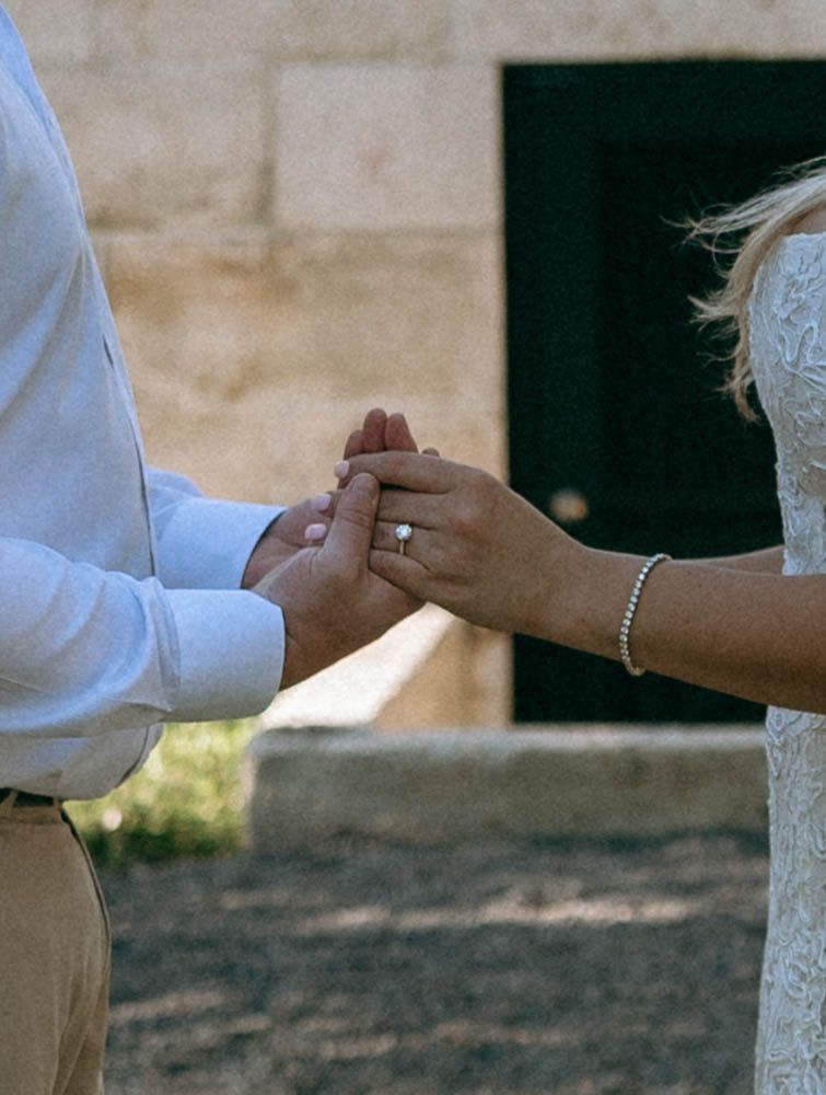 Couple holding hands during wedding ceremony, bride wearing diamond engagement ring and bracelet, groom in light blue shirt.