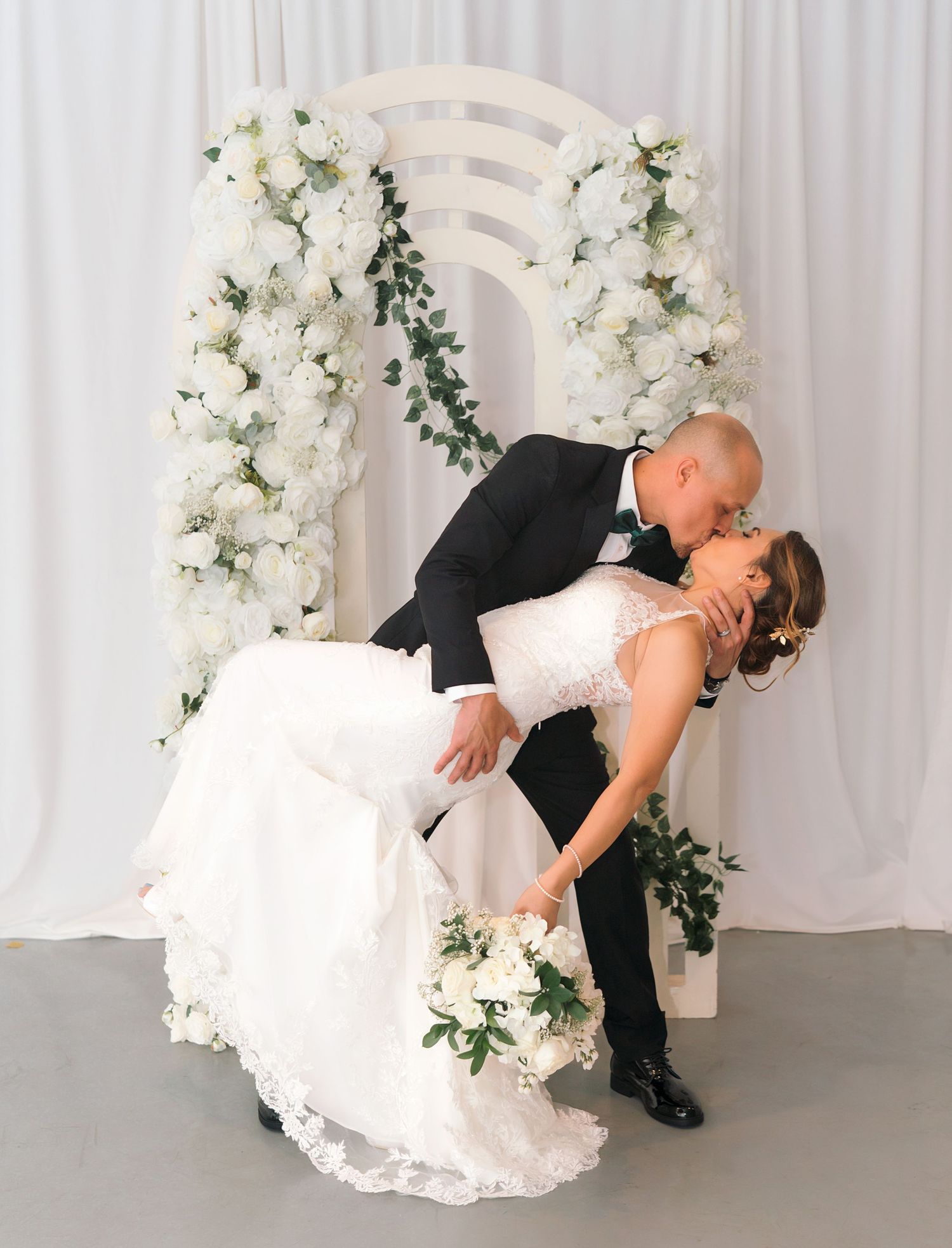 Romantic wedding photo series showing a couple in formal attire dipping pose against white floral backdrop.