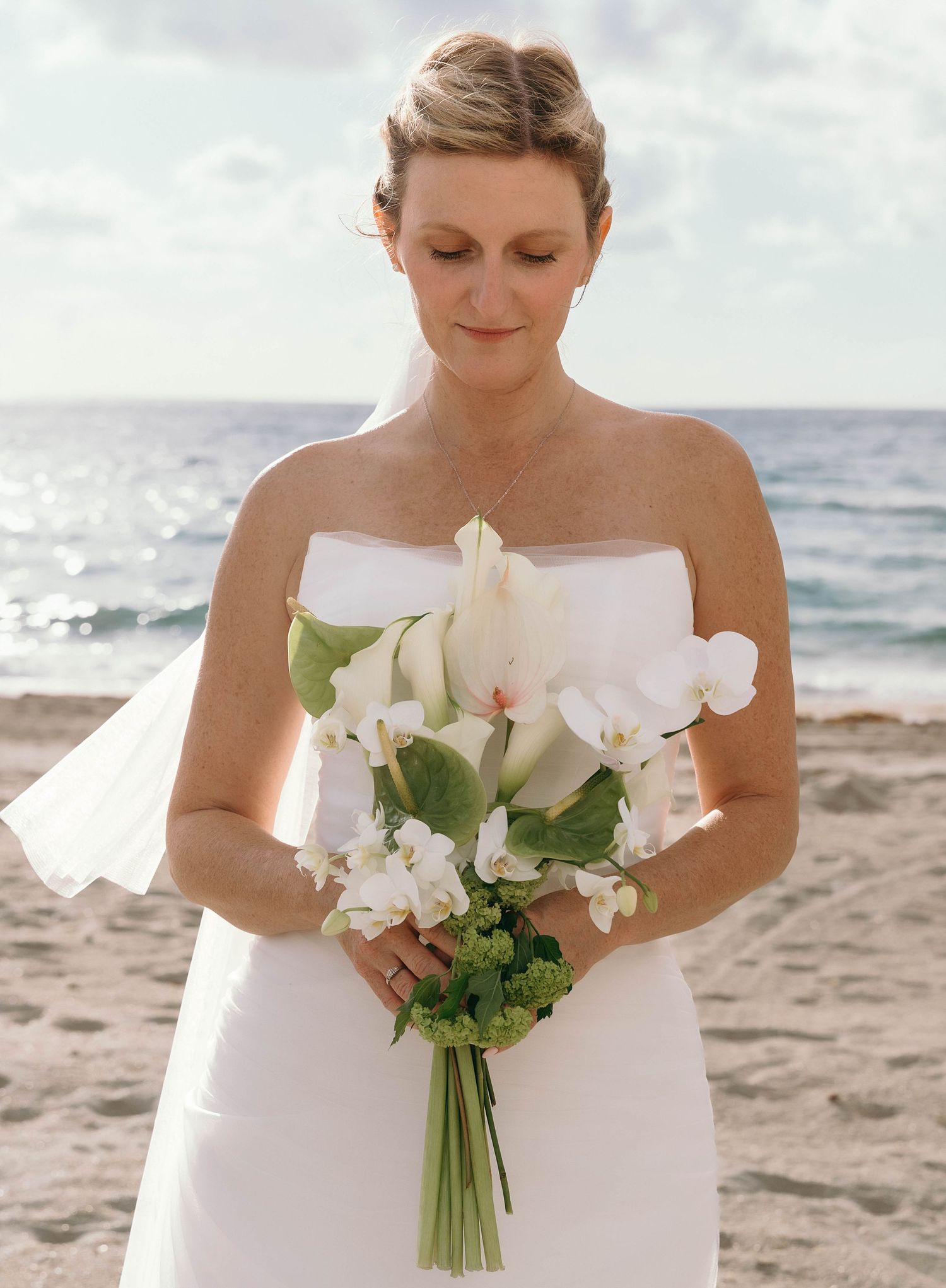 Bride holding white floral bouquet on beach with ocean view.