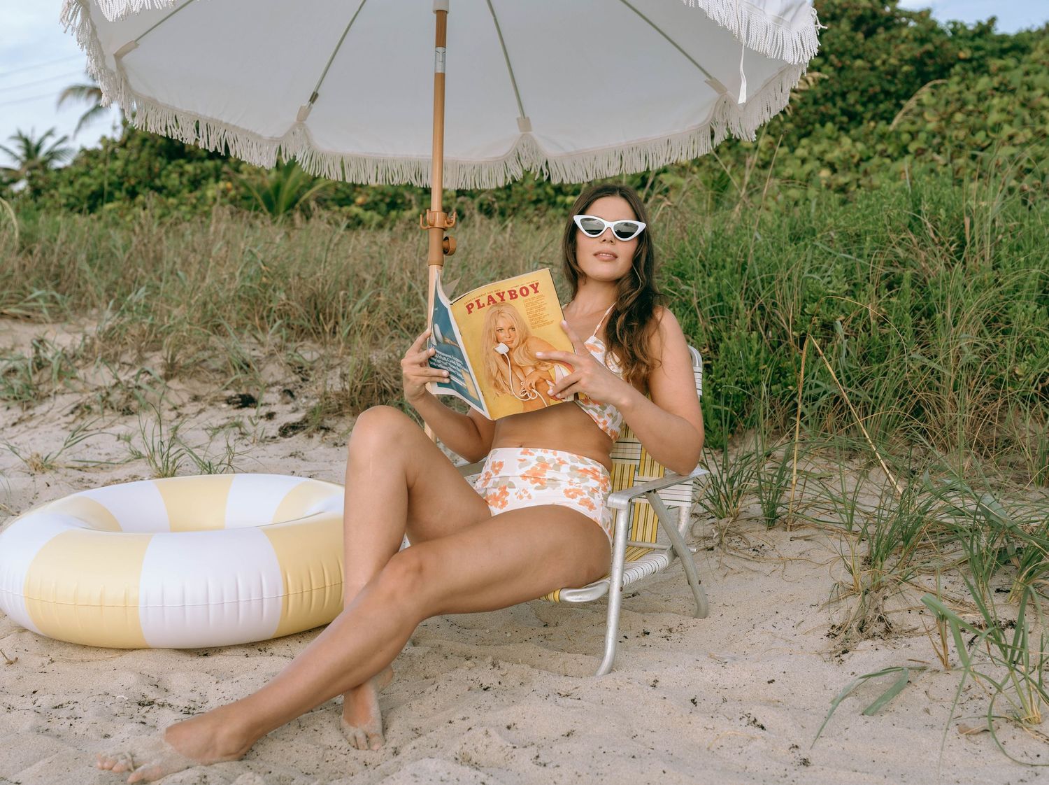 Woman reading a magazine on a beach chair under a fringed umbrella with a pool float nearby.