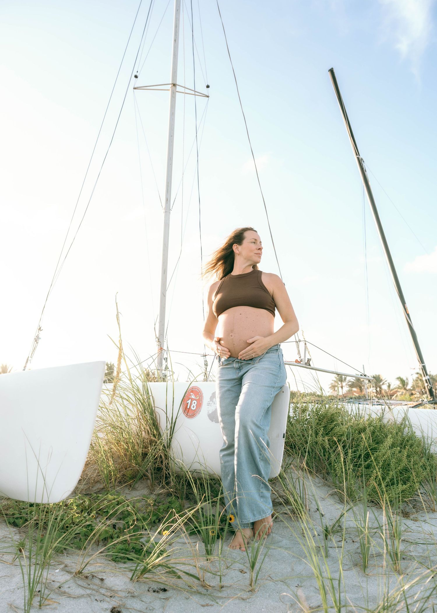 Pregnant woman in brown crop top and jeans standing barefoot on sandy beach beside white sailboat.