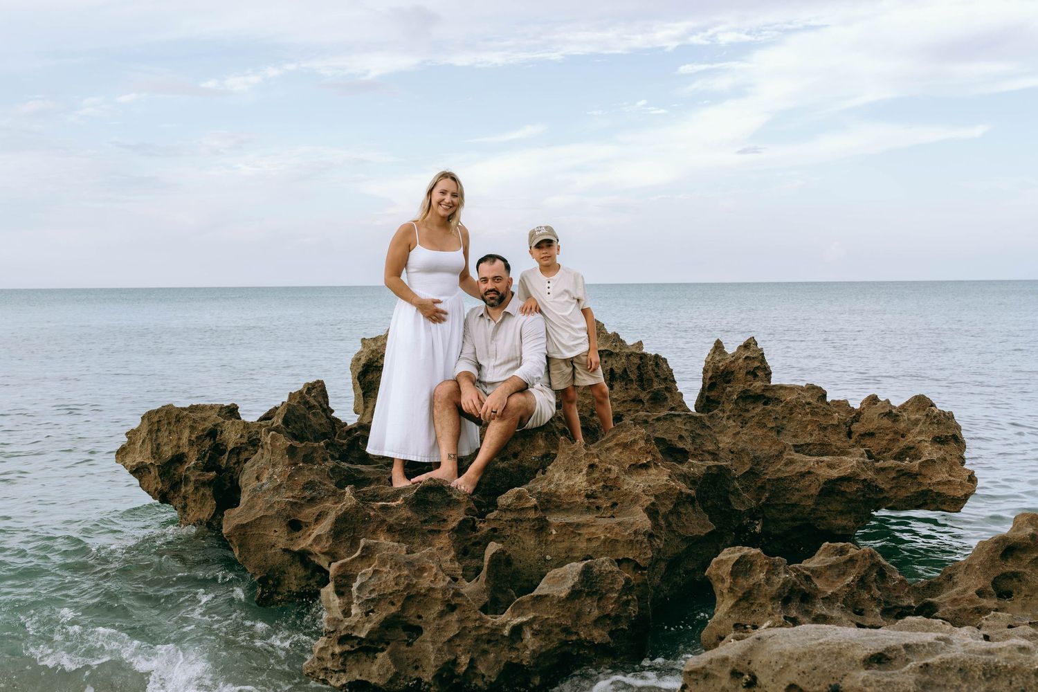 Family of three posing on rocky shoreline with ocean waves and cloudy sky backdrop.