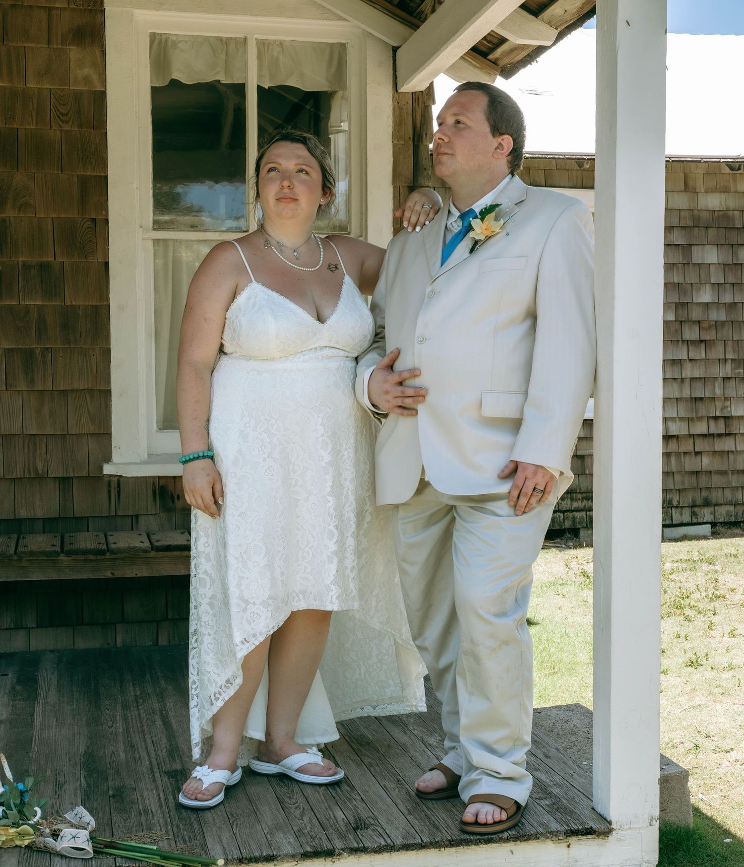 Wedding couple poses on rustic wooden porch, bride in white lace dress and groom in tan suit with floral boutonniere.