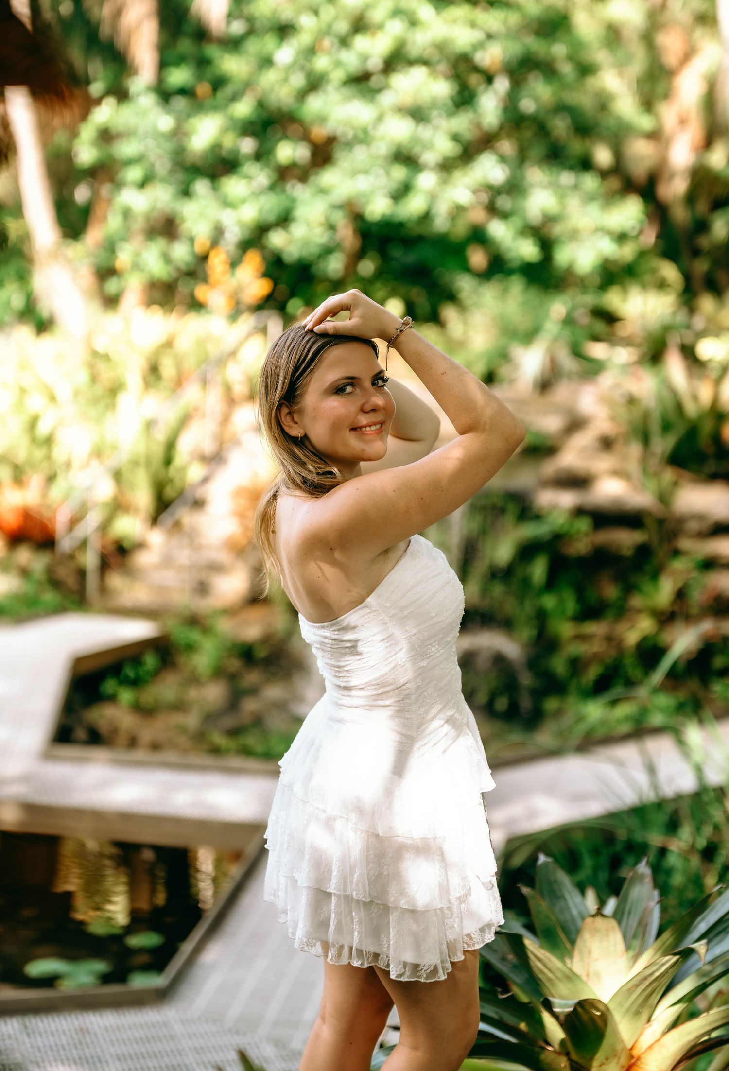 Woman in white ruffled mini dress posing in a lush tropical garden near a reflective pond.