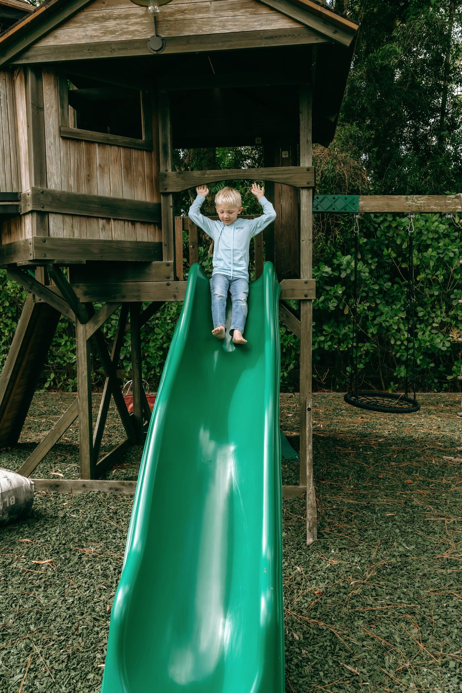 Young child sliding down a green slide at a wooden treehouse playground outdoors.