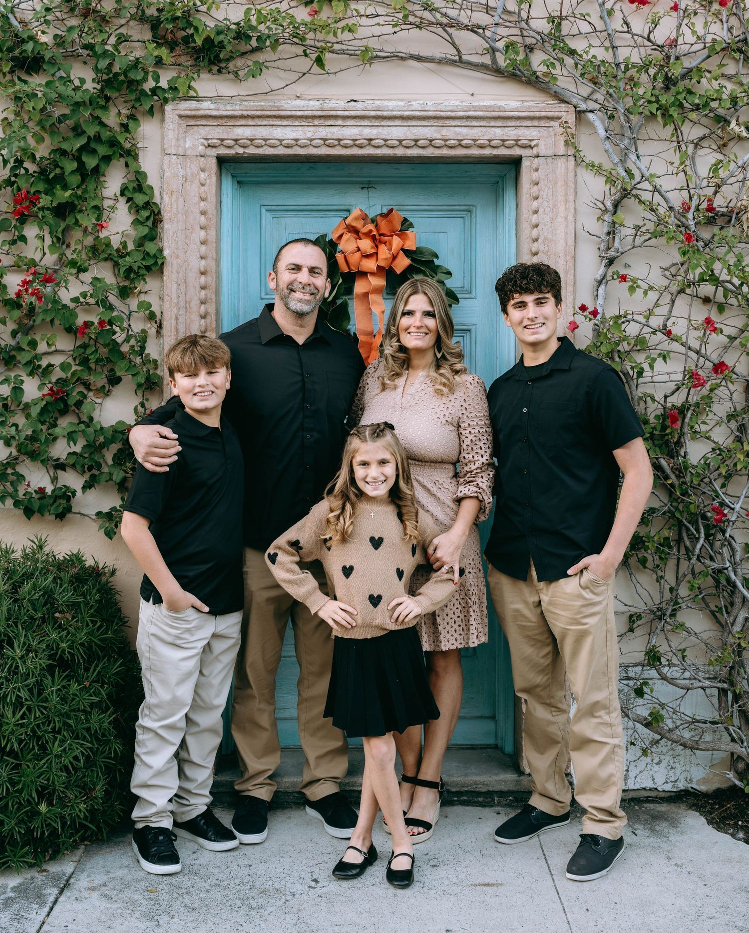 Family of five posing happily in front of a teal door decorated with an orange bow wreath, surrounded by ivy.
