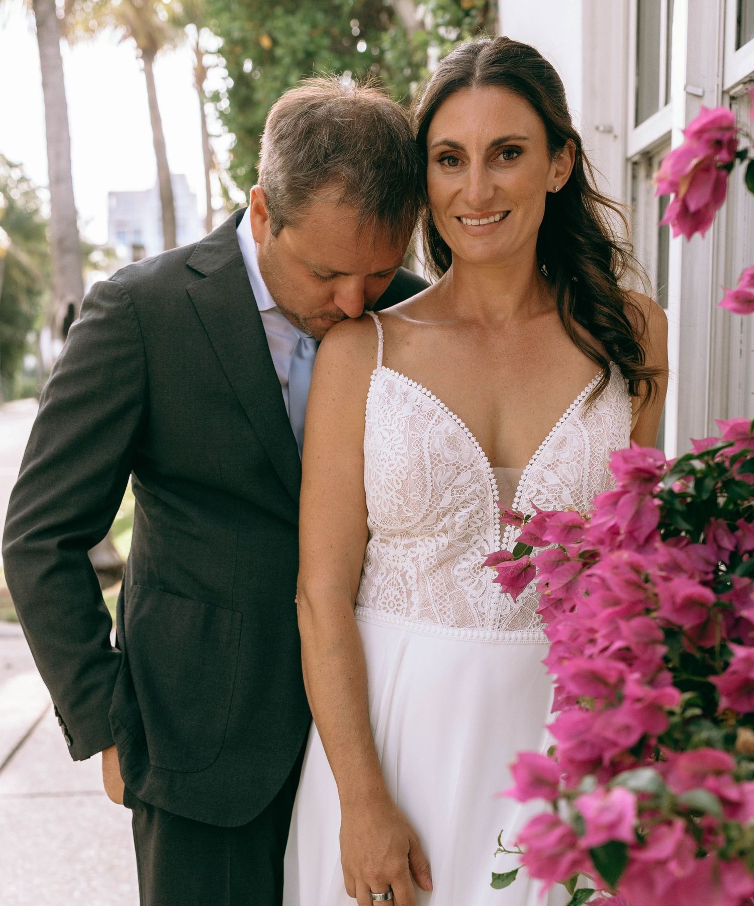 Bride in lace wedding gown with groom in gray suit posing near pink bougainvillea flowers outdoors.