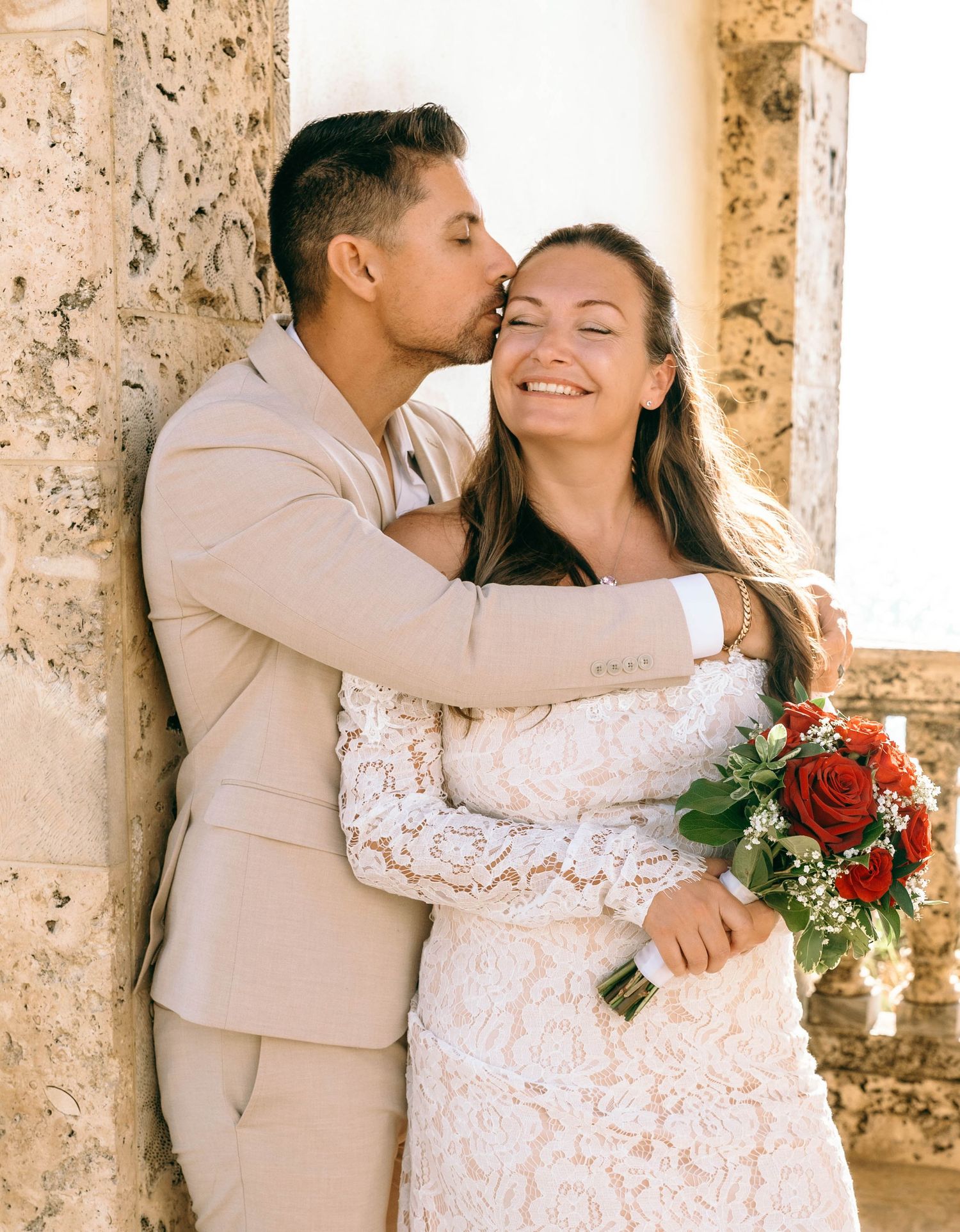 Groom in beige suit kissing bride in lace dress holding red roses bouquet outdoors.