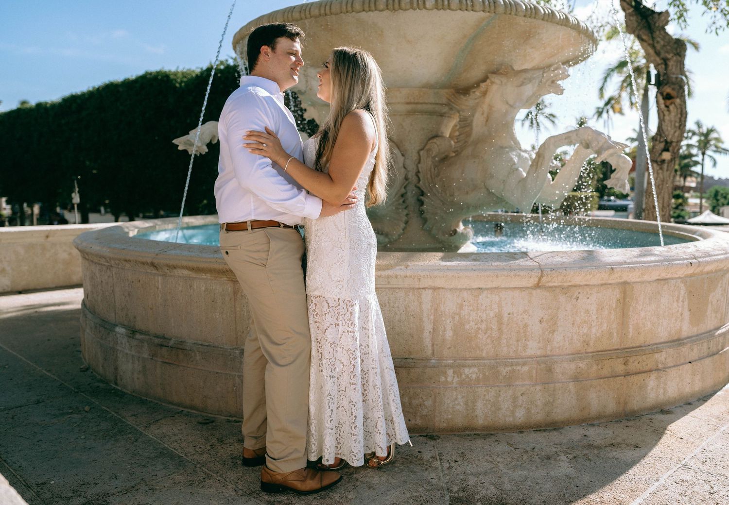 Couple embracing romantically in front of an elegant stone fountain, woman in white lace dress, man in khaki pants.