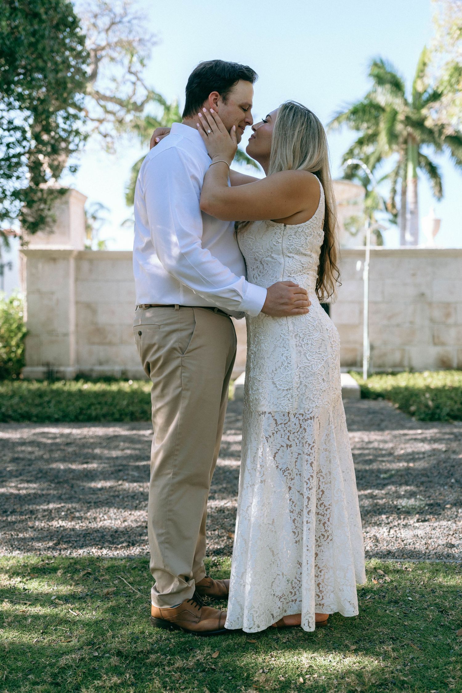 Couple sharing a romantic kiss in a lush garden, man in white shirt and khakis, woman in white lace dress.