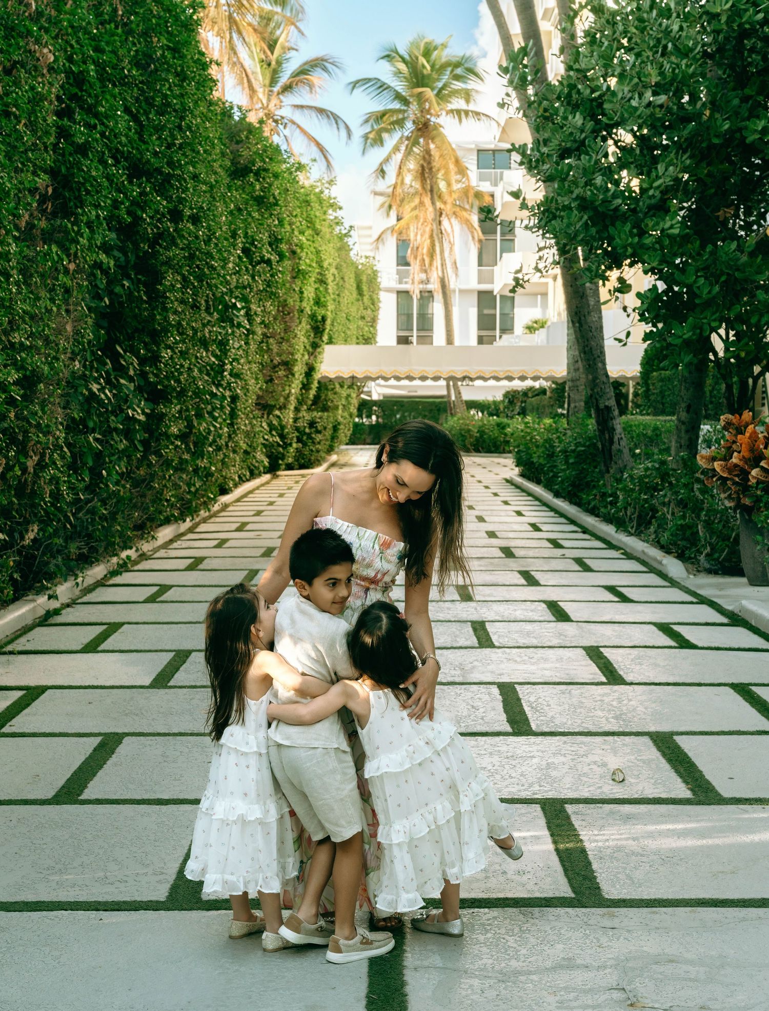 Mother in white dress hugs three children on elegant palm tree-lined garden pathway. Worth Avenue, Palm Beach, FL