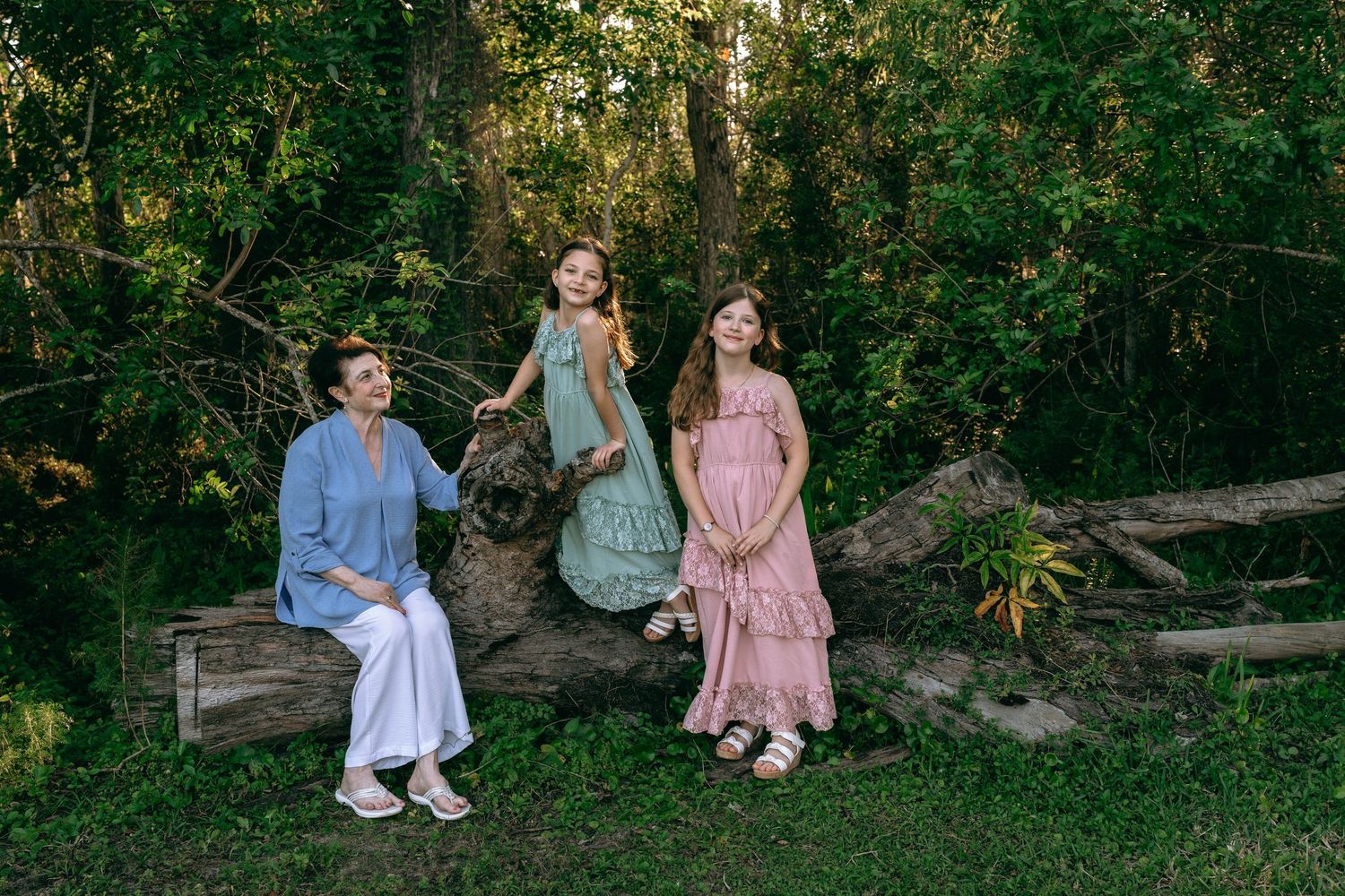 Three females in pastel dresses pose on a fallen log surrounded by lush green forest trees. Colt Farms