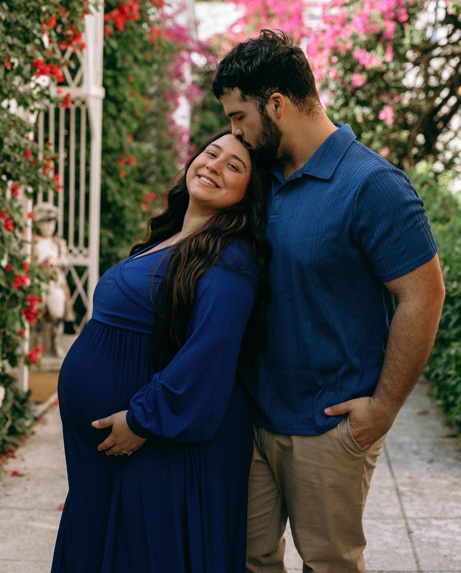 Happy pregnant couple in blue outfits embracing in a colorful bougainvillea-lined garden path. Worth Avenue, Palm Beach, FL