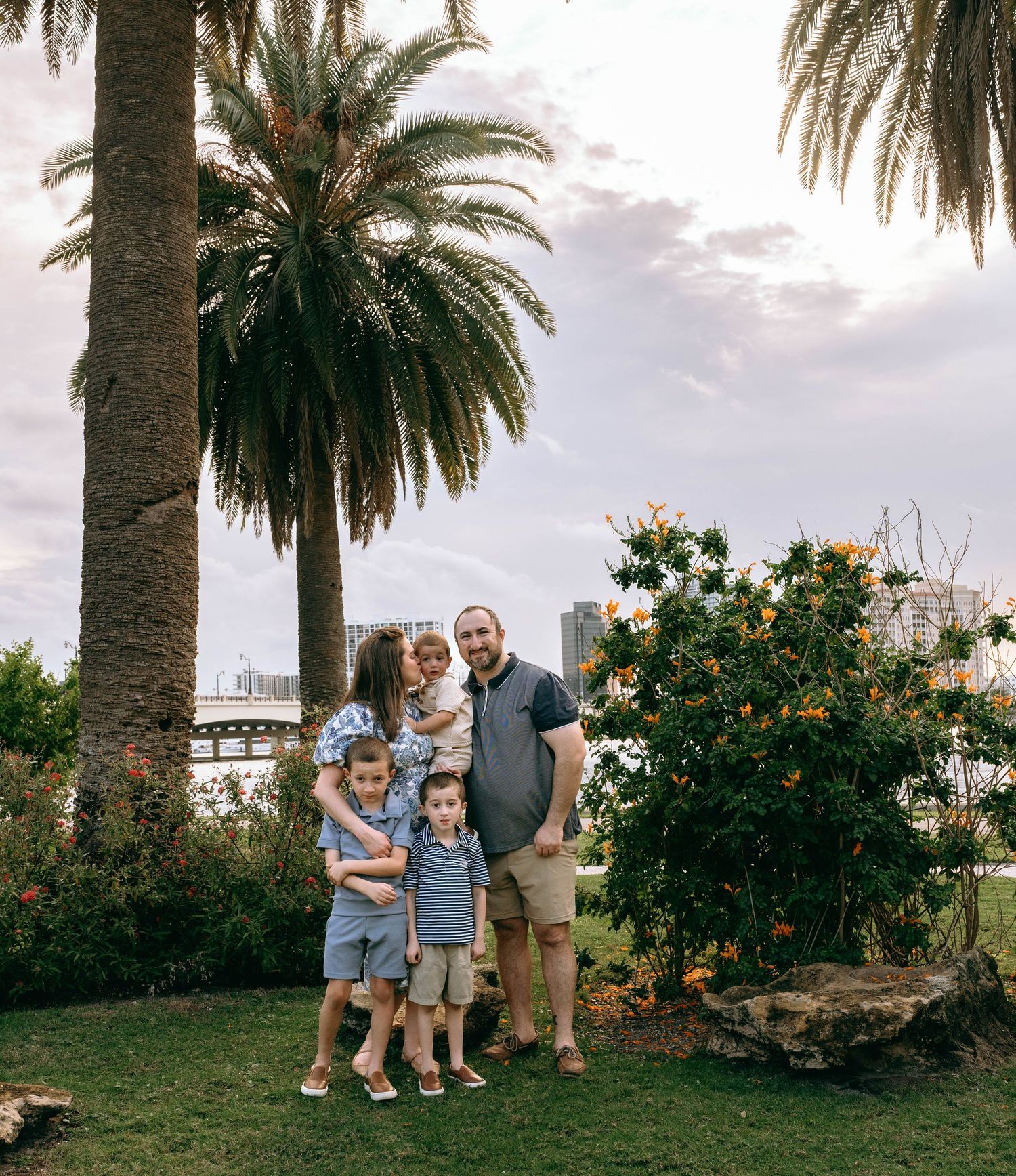 Family of five posing together in tropical garden with palm trees and city skyline in background.  Family photos at Bradley Park Palm Beach,